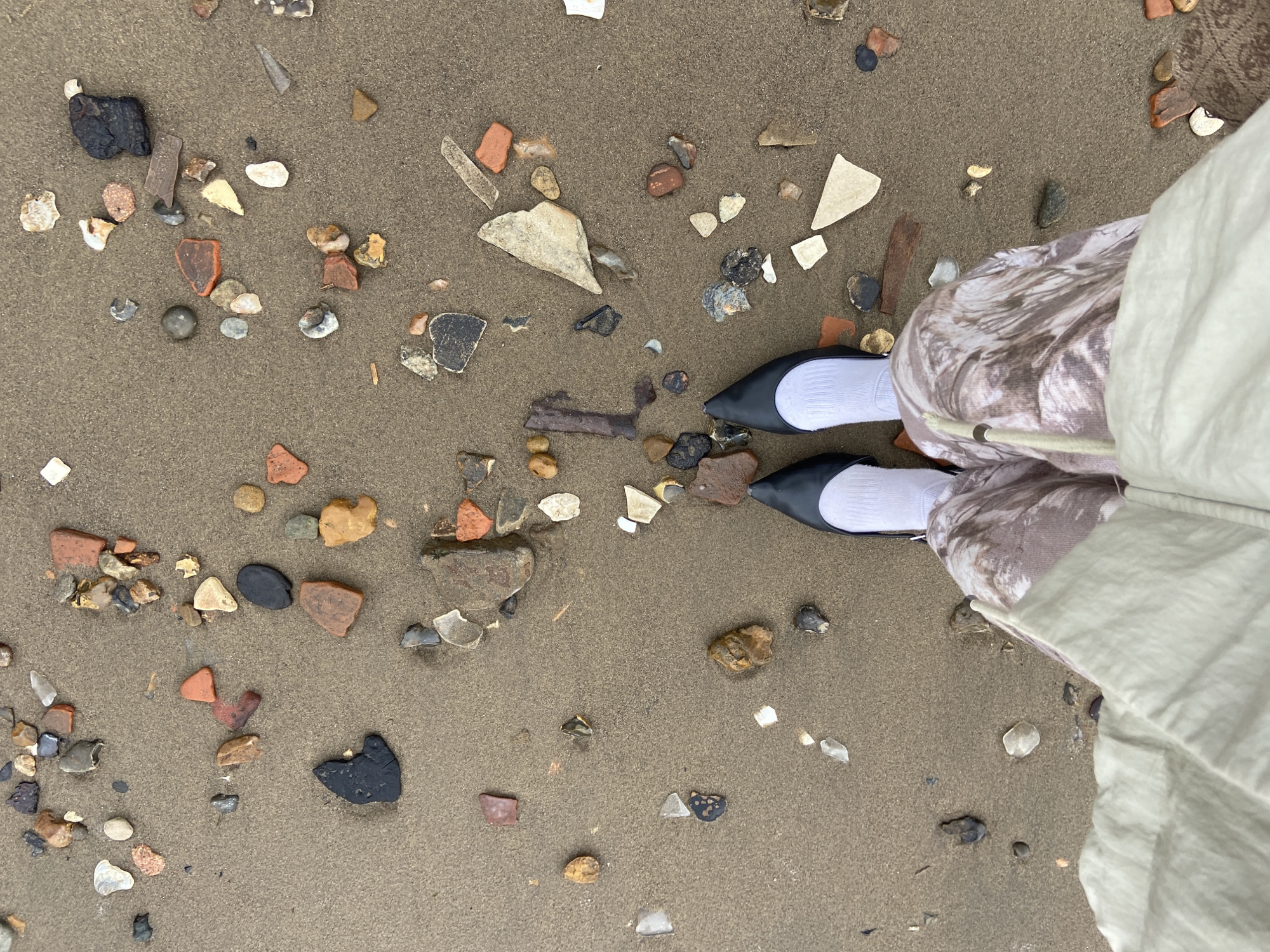 The image depicts a sandy beach littered with small stones, pebbles, and shards of pottery and glass. A person is standing on the beach, wearing black pointed-toe flats with white socks, patterned trousers, and a light-colored jacket. The perspective is from slightly above, looking down. The scene suggests a beach environment, potentially urban given the presence of pottery shards. There is no explicit information identifying the location as London.