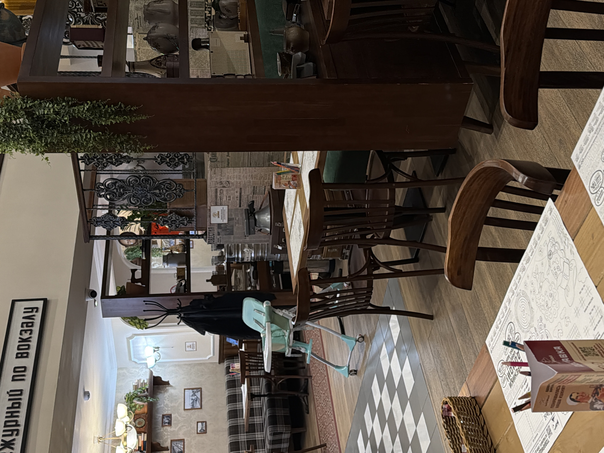 The image captures the interior of a restaurant or cafe, likely in Moscow, Russia, given the visible Cyrillic text. The scene presents a well-appointed dining area with a blend of classic and rustic decor.
In the foreground, a wooden table holds a paper placemat designed for coloring, accompanied by a box of colored pencils labeled "ЗАБАВКА" (Zabavka, meaning "toy" or "fun") and a wicker basket. A light blue high chair for a child is positioned next to this table, suggesting a family-friendly atmosphere. The floor features a combination of light wood planks and a distinctive checkerboard pattern of gray and white tiles.
The mid-ground reveals more seating, including wooden chairs and a booth with plaid upholstery. A tall, dark wooden shelving unit acts as a partial room divider, decorated with various items such as vases, a globe, and cascading green artificial plants. Behind this unit, the wall is covered in wallpaper designed to look like old newspapers, with headlines including "ИЗВЕСТИЯ" (Izvestia), a well-known Russian newspaper.
Further into the background, additional tables and chairs are visible. The lighting comes from elegant chandeliers and wall sconces, illuminating framed pictures on the walls. A prominent black and white sign on the left wall reads "ДЕЖУРНЫЙ ПО ВОКЗАЛУ" (Dezhurnyy po Vokzalu), which translates to "Station Duty Officer" or "Duty Officer at the Station," hinting at a possible railway station theme or location. Small security cameras are mounted on the ceiling. The establishment appears clean and organized, ready for customers, though no people are currently visible in the shot. The time of day is not discernible, as the indoor lighting provides the main illumination.