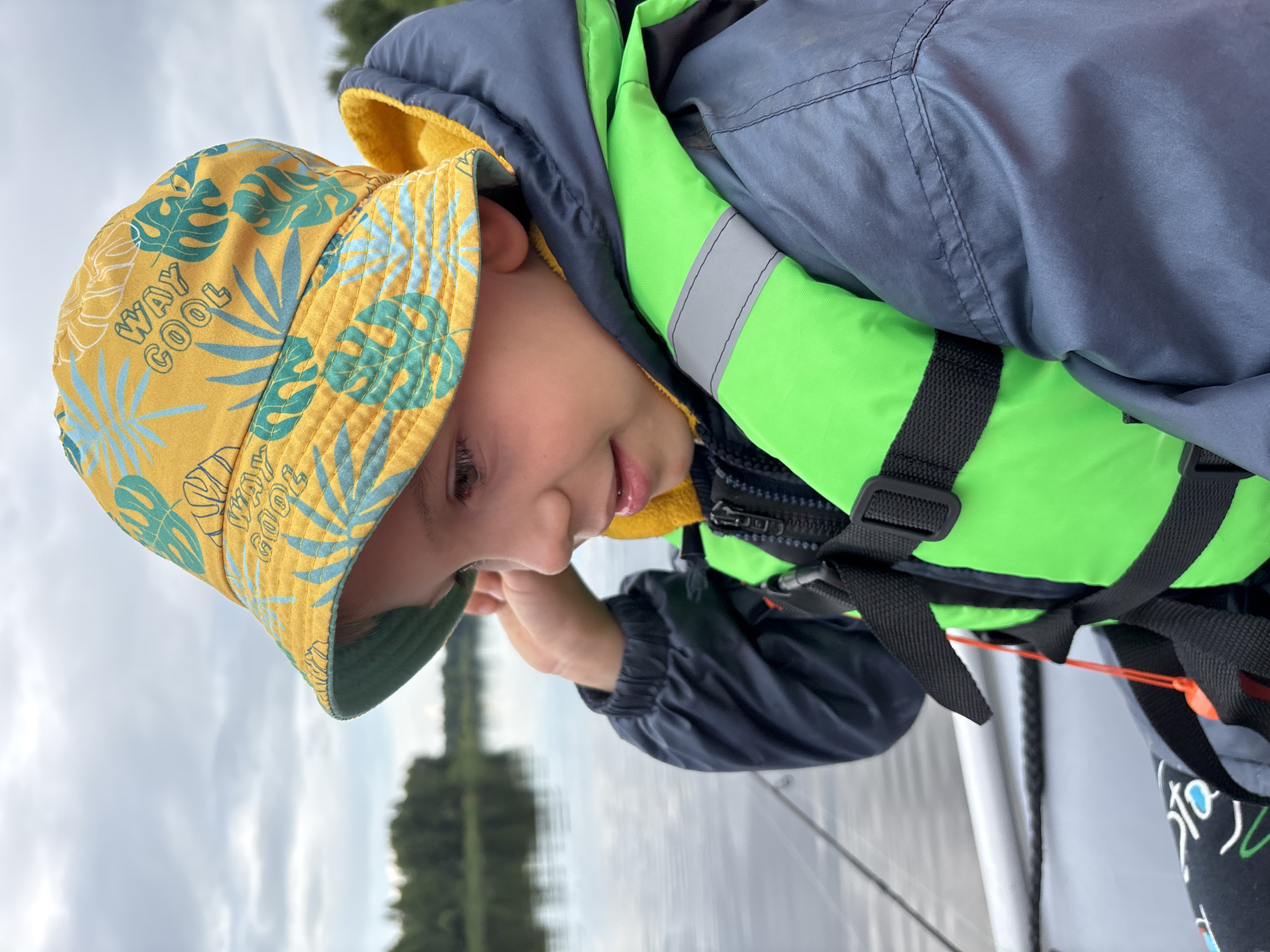 A young boy, wearing a yellow bucket hat with a tropical leaf print and the words "WAY COOL" printed on it, and a bright green life vest over a dark blue jacket, is shown from the chest up. He is looking down and to the left, with a slight smile on his face. His right hand is near his cheek. The background shows a body of water with trees on the distant shore, under an overcast sky. The scene appears to be outdoors, possibly on a boat, given the presence of what looks like a railing or edge of a vessel. The lighting suggests it is daytime.