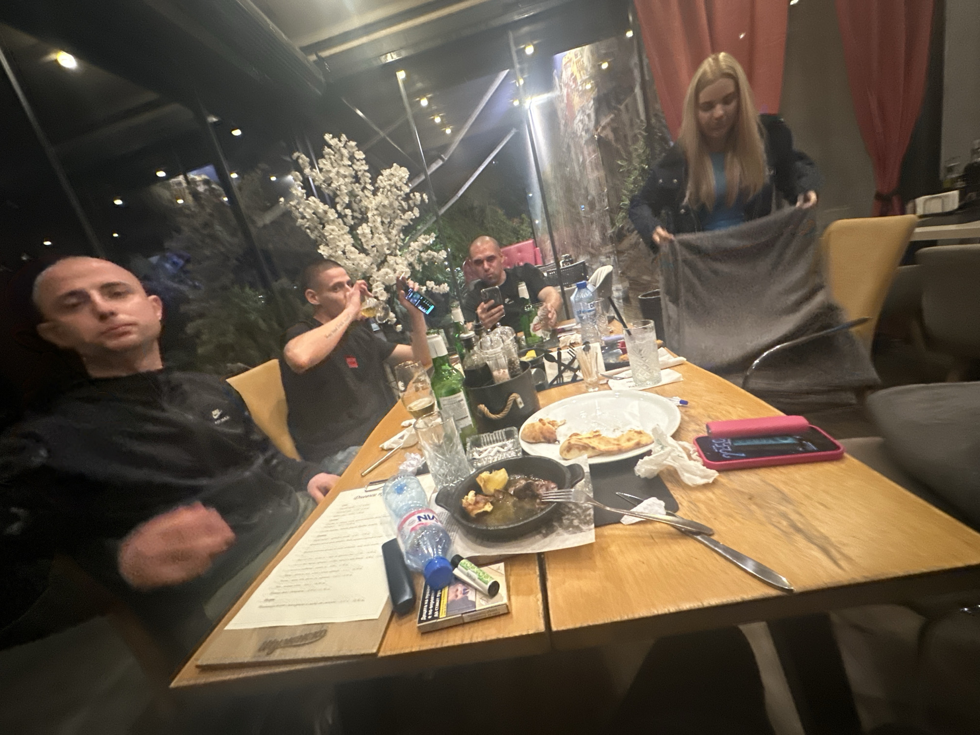 The image shows four men and one woman seated around a table in what appears to be a restaurant or bar setting. The location is Sofia, Bulgaria, as indicated by the Cyrillic writing on the cigarette pack on the table.
The men are dressed in casual clothing. One is looking directly at the camera. A second man is holding up a cell phone, possibly taking a picture. A third man is looking at his cell phone. The woman is holding up a grey blanket.
The table is covered with plates, glasses, bottles, food remnants, a phone in a pink case, a bottle of water, silverware, a cigarette pack, a vape pen and a menu.
The restaurant has large windows overlooking an exterior area with trees. The interior decor includes modern lighting and decorative flowers. The atmosphere suggests an evening gathering.