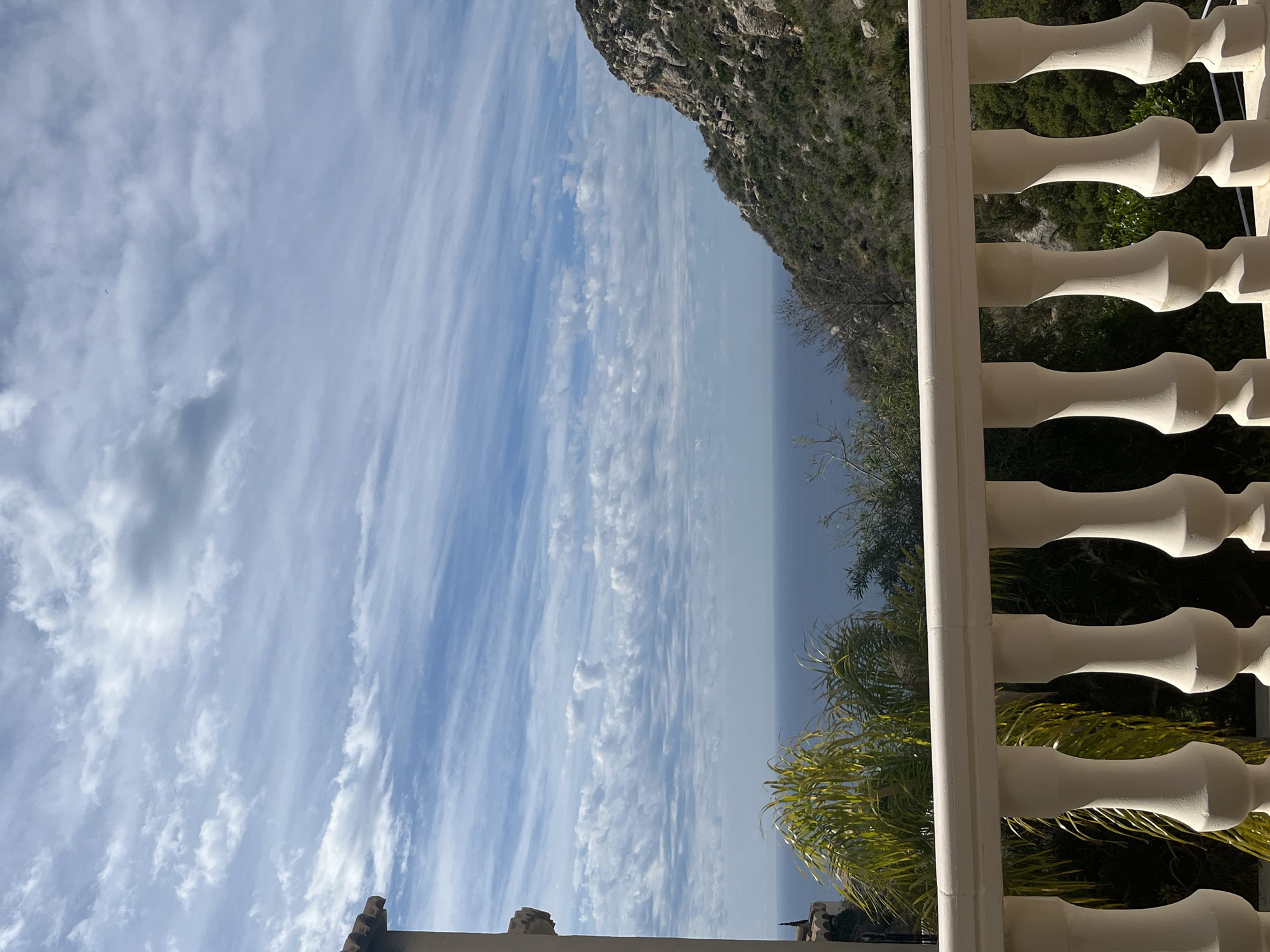 From a balcony in el Poble Nou de Benitatxell / Benitachell, Spain, a view encompasses a partly cloudy sky, the sea, and a rocky cliff. A white balustrade runs along the right side of the frame, partially obscuring a green palm tree and other vegetation. The sky displays elongated cloud formations. A rocky cliff borders the horizon on the upper right. The sea is a muted blue in the distance. The scene appears to be captured during daylight.