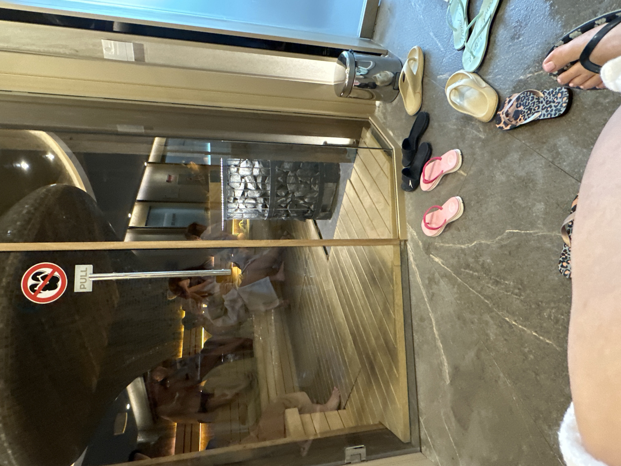 This image captures a view into a sauna from the outside, taken at a spa or wellness center in Kazanlak, Bulgaria. Inside the glass-fronted sauna, several individuals are visible, relaxing on wooden benches. Their forms are partially obscured by reflections on the glass. A prominent sauna heater, filled with rocks, stands against the far wall inside. The interior of the sauna features warm-toned wooden panels.
Outside the sauna, on a dark grey tiled floor with lighter marbling, a collection of flip-flops of various colors – including beige, black, pink, light green, and a leopard print pair – are scattered near the entrance. The legs and feet of a person wearing a leopard-print flip-flop are visible in the foreground, suggesting the perspective of someone standing or sitting just outside the sauna. A chrome-colored metal container is mounted on the wall to the left of the door.
Visible text on the glass door includes a red circular sign with a crossed-out symbol resembling a pair of flip-flops or shoes, indicating that footwear should not be worn inside. Adjacent to this sign, a white label with the word "PULL" is affixed near the door handle. The scene conveys a calm and relaxing atmosphere typical of a spa environment. The lighting suggests an indoor setting, likely during the daytime or a well-lit interior.