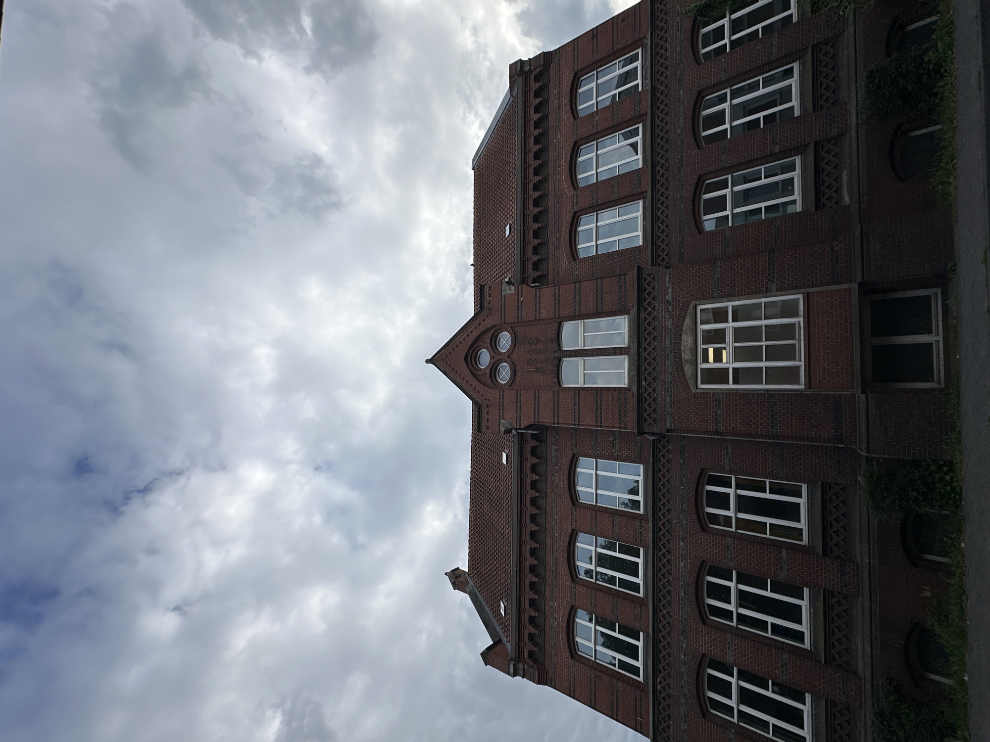 A low-angle exterior photograph depicts a multi-story institutional building, likely a school, constructed of dark red brick with white-framed windows, situated under an overcast sky in Stade, Germany. A central gable prominently features the year "1888" in brickwork beneath three circular windows. The building is topped with a red tiled roof, featuring small skylights and a chimney-like structure on its left side. Decorative brick patterns, including a zigzag frieze and dentil-like elements, are visible below the upper-floor windows and along the eaves. The facade displays numerous tall, narrow windows, some with arched tops, others rectangular; one lower central window emits light from within. Sparse green vegetation, primarily ivy, grows along the base of the building, partially obscuring small arched ground-level openings. The sky is uniformly grey with heavy cloud cover, providing diffuse lighting. No individuals or overt activities are visible in the scene.