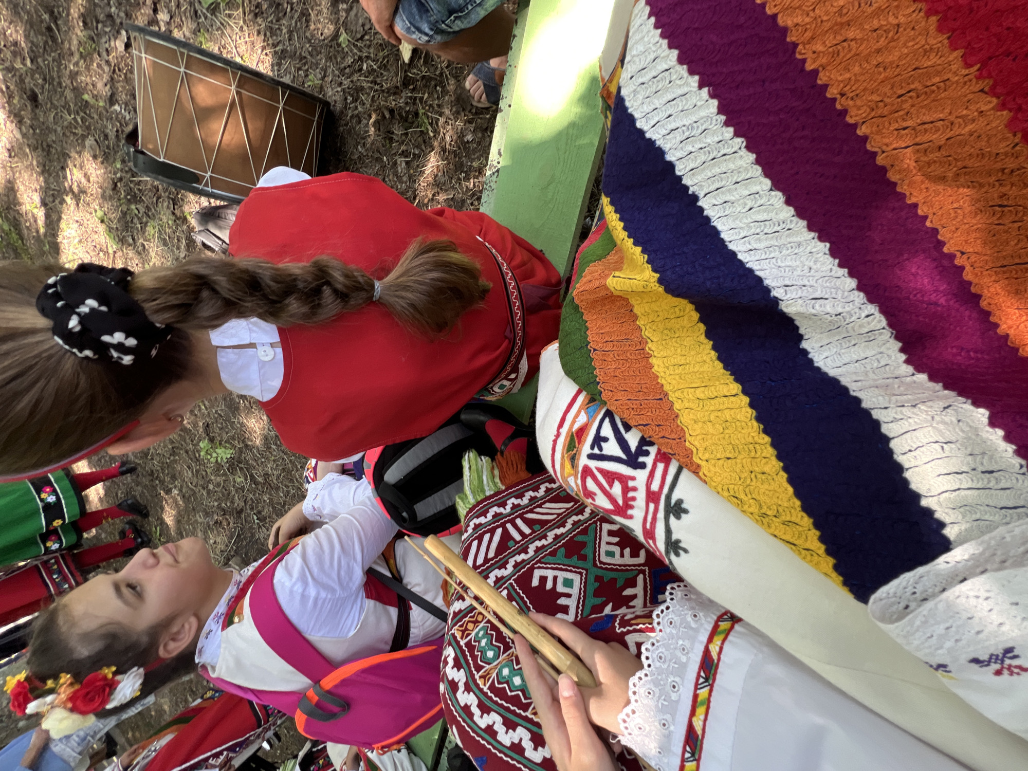The image depicts an outdoor cultural gathering in Bolyarovo, Bulgaria. Two young females, dressed in traditional Bulgarian folk costumes, are prominent in the foreground and midground.
The girl on the left, viewed in profile, wears a white embroidered blouse with lace trim, a light-colored vest, and has her dark hair pulled back and adorned with red, yellow, and white fabric flowers. A bright pink backpack with orange straps is worn over her costume. She is seated, looking towards the right with a serene expression.
The girl on the right, viewed from behind, has her brown hair styled in a long braid, secured at the top with a black and white polka-dotted scrunchie and at the end with a grey elastic band. She wears a red sleeveless garment or pinafore over a white collared shirt, with a patterned belt featuring geometric designs around her waist. She is seated on a green wooden bench, part of which is visible beneath her.
In the bottom left foreground, a pair of hands with a white, lace-trimmed sleeve holds a light wooden stick-like instrument, possibly a shepherd's flute (kaval) or similar, which rests on a richly embroidered red textile featuring geometric patterns in white, green, and blue. Adjacent to this is a folded, multi-colored striped textile, likely a blanket or shawl, displaying bands of yellow, navy blue, white, magenta, orange, and red.
Behind the girl on the right, a traditional wooden frame drum (likely a davul or tapan) with visible rope lacing is lying on the uneven, earthen ground. The ground itself is covered with dry leaves and sparse foliage, indicative of an outdoor, possibly wooded, setting. A partial view of a person's leg in blue jeans and brown sandals is visible to the right of the girl on the bench. In the blurred background, other individuals are partially visible, some also in traditional folk attire, including red and green skirts with white embroidery, suggesting a larger assembly or performance. The scene is illuminated by natural daylight.