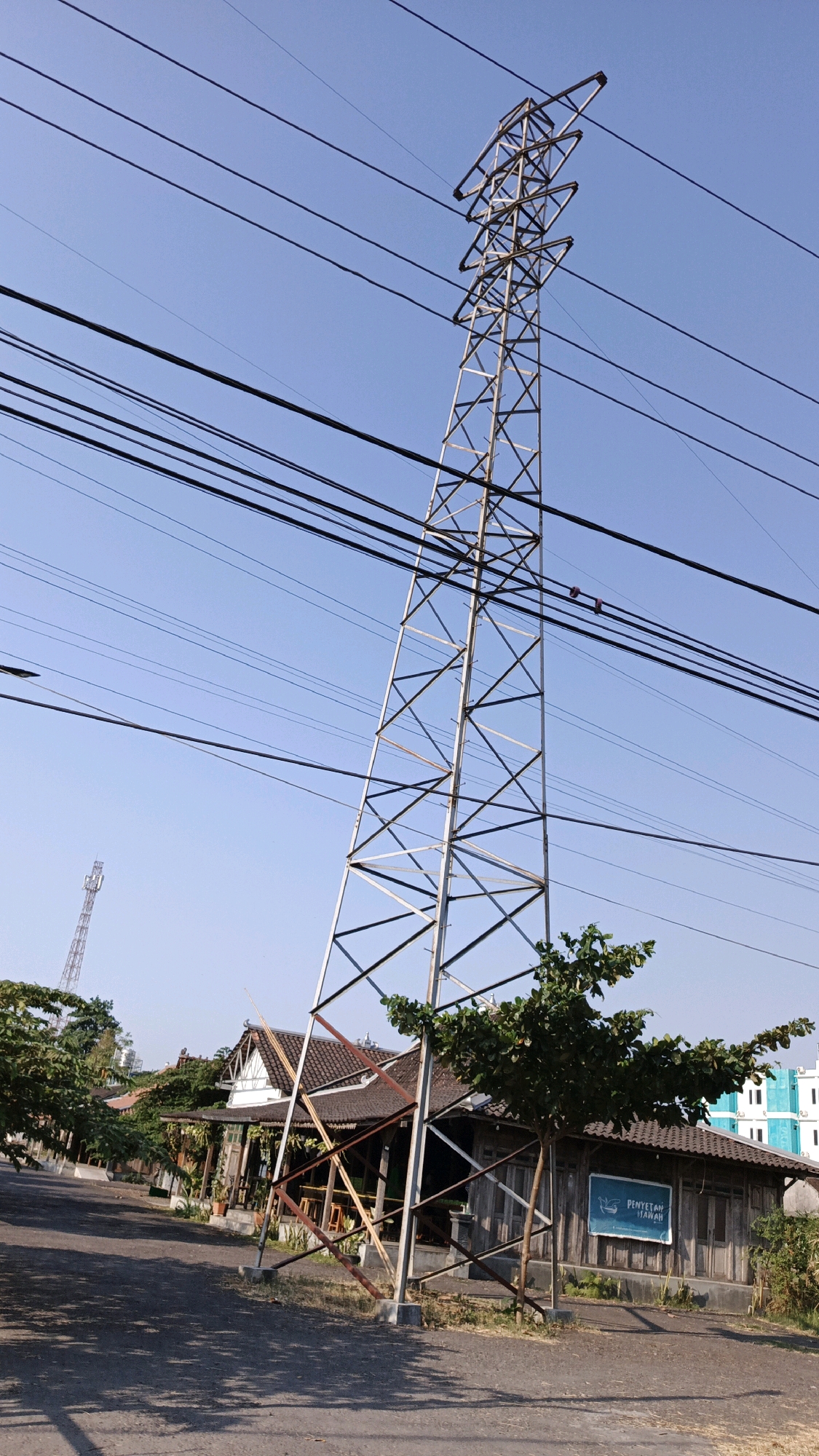 This is a photograph taken in Pabelan, Indonesia, capturing a scene with a tall, industrial metal tower supporting numerous electrical wires against a clear blue sky. The tower is angled significantly to the right, giving the impression of instability. In the foreground, a paved road is visible, with shadows cast by trees and the tower. To the left of the tower, a building with a traditional-looking tiled roof and wooden walls stands, with some greenery around it. A blue sign with white text is mounted on the wall of this building, reading "PENYETAN UMAH". Further in the background, more buildings and trees are present. In the distance, another tower, likely a telecommunications tower, is visible on the left side of the frame. The overall environment suggests an outdoor, possibly rural or semi-urban setting, under daylight conditions.
