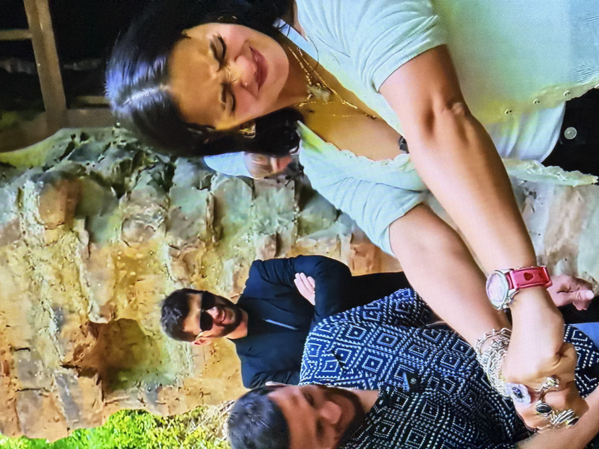 In an outdoor setting with a stone wall and greenery, three individuals are visible. In the foreground, a woman with dark hair is shown from the chest up, her face contorted in a grimace as she clutches her arm. She is wearing a light-colored, open-front cardigan over a white top, multiple necklaces, and several rings on her left hand. A man in a black suit and sunglasses stands behind her with his arms crossed, smiling. To the left, another man wearing a black and white patterned shirt is also looking down and smiling. The focus of the image is on the woman and her interaction with an unseen object or person, as indicated by her strained expression and the way she is holding her arm.