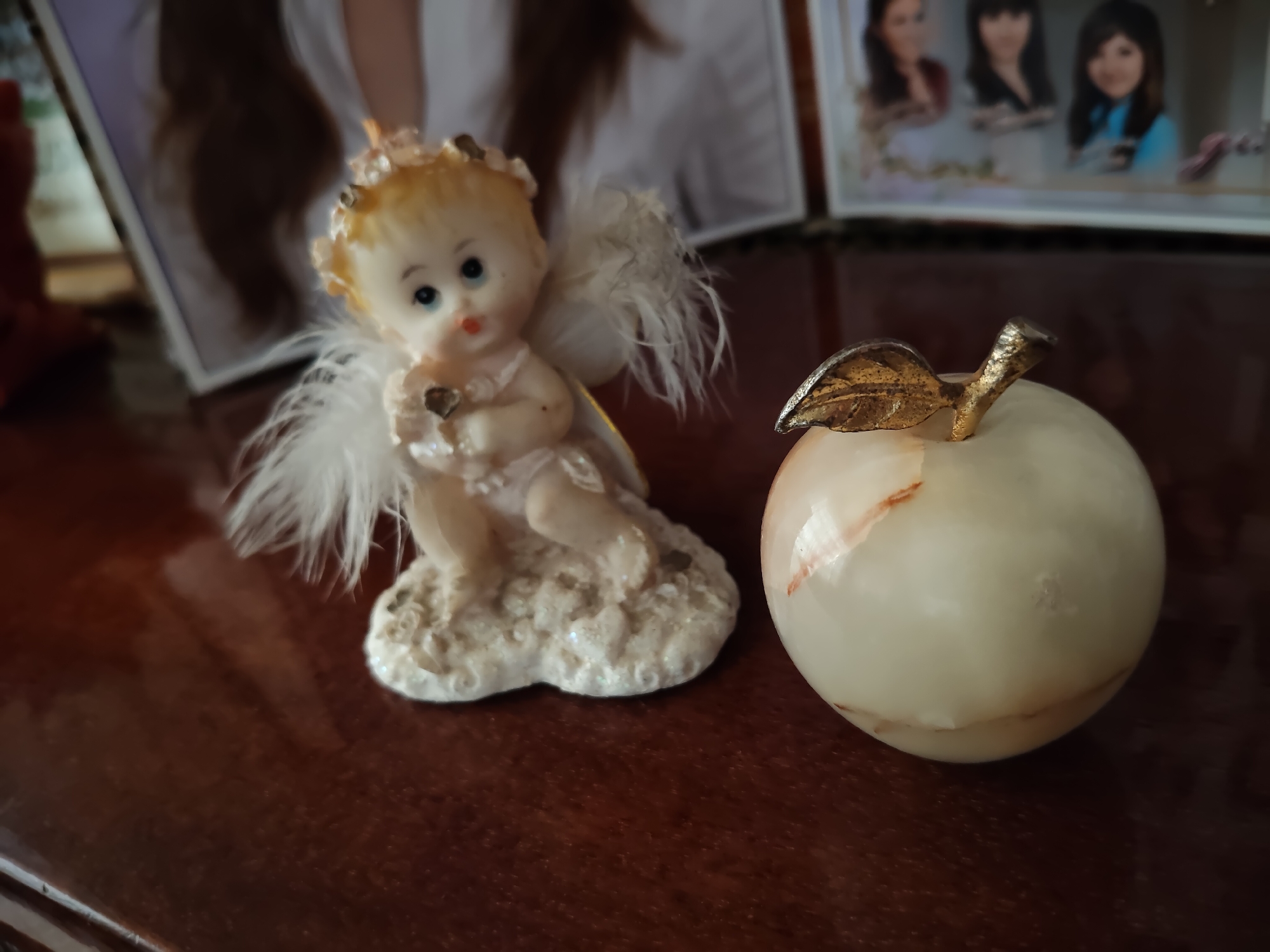 This is a close-up, eye-level shot of two decorative objects placed on a polished wooden surface. In the foreground, to the left, is a small figurine of a baby angel with blonde hair, white feathered wings, and a halo. The angel is sitting on a textured base and holding a small heart. To the right of the angel is a round, off-white decorative apple with a metallic leaf and stem. In the background, out of focus, are framed photographs. The overall scene is dimly lit, with the objects catching the light. The environment is likely a home interior, possibly a shelf or table in Gîrbova, Moldova.