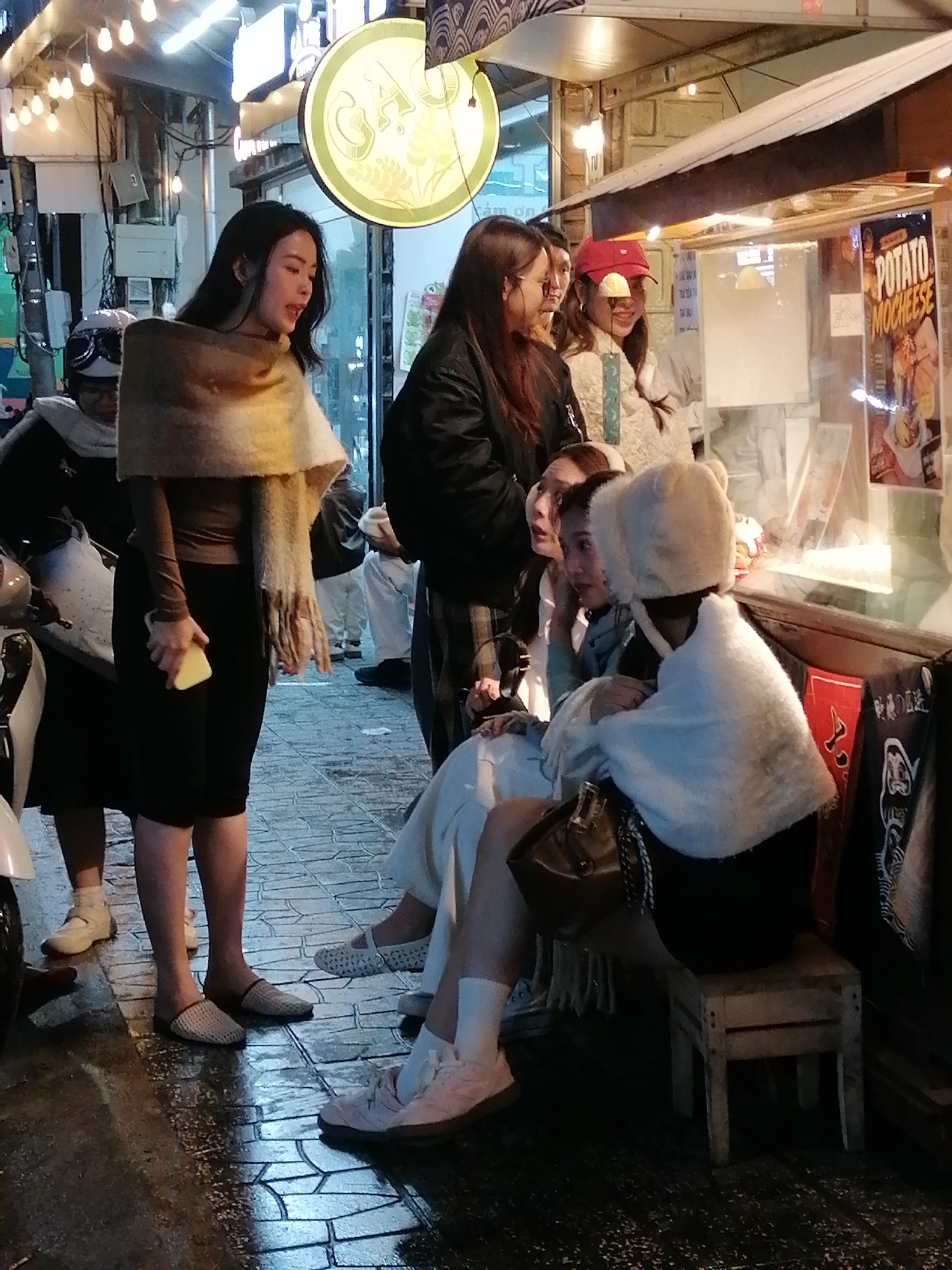 The image depicts a group of young women gathered around a food stall on a wet, tiled street in Da Lat, Vietnam. The lighting suggests it is evening or night. A woman in the foreground, wearing a brown top and black skirt with a fringed scarf, holds a yellow triangular object, possibly food. She is looking to her left. To her right, another woman is seated, wearing a white furry hat and wrapping, facing away from the camera. Behind them, other individuals are visible, some interacting with the stall which displays what appears to be food items and signage, including one that says "POTATO MOCHEESE". The overall atmosphere is casual and communal, with people gathered at a street-side vendor.