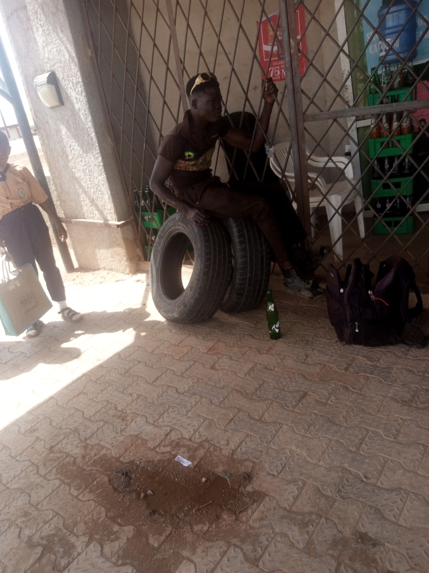 Two young men, one wearing sunglasses, sit on tires outdoors in front of a metal grate. The man on the left wears a brown t-shirt and dark pants. The man on the right wears a dark t-shirt and dark pants. A green bottle lies on the ground in front of them, and a black backpack rests beside the bottle. To the left, a person in a yellow shirt and dark pants carries a shopping bag. The ground is paved with interlocking bricks, and there is a wet patch on the ground in the foreground. Behind the men, crates of bottled drinks are visible through the grate, as are chairs and a water cooler.