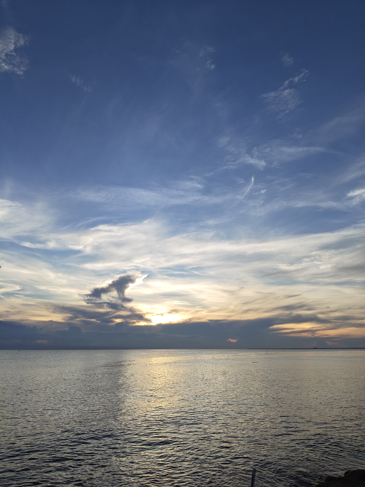 The image shows a tranquil seascape under a partly cloudy sky. The sky transitions from a deep blue at the top to a lighter, more diffused blue near the horizon, where it mingles with patches of white and gray clouds. The sun, partially obscured by a layer of clouds, casts a golden glow on the water's surface. The sea itself is calm, with gentle ripples visible across its expanse. The sunlight reflects off the water, creating a shimmering effect.
The location is likely in Lhokseumawe, Indonesia, as stated. The view is an open, natural scene without buildings, street furniture, or any specific identifying markers of an urban area. The picture captures a moment of natural beauty and tranquility. There are no people or other subjects visible.