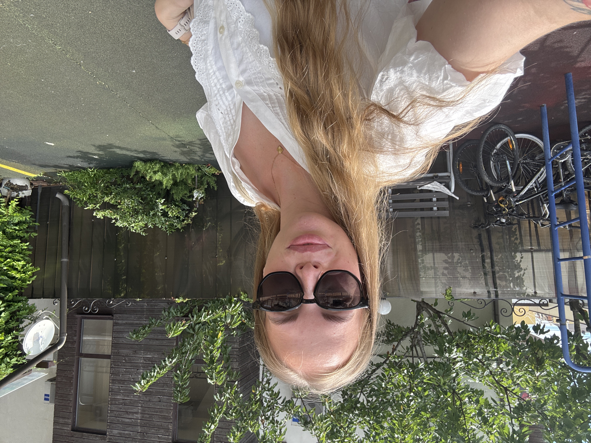 A young woman with blonde hair and sunglasses is taking a selfie outdoors. She is wearing a white, lace-trimmed blouse and a gold necklace. In the background, there are trees, a wooden fence, a building with windows, bicycles, and a blue metal structure. The ground appears to be a dark, textured surface, possibly asphalt or rubberized playground material. The scene is illuminated by natural daylight, suggesting it is daytime. The location is identified as Sirius, Russia.