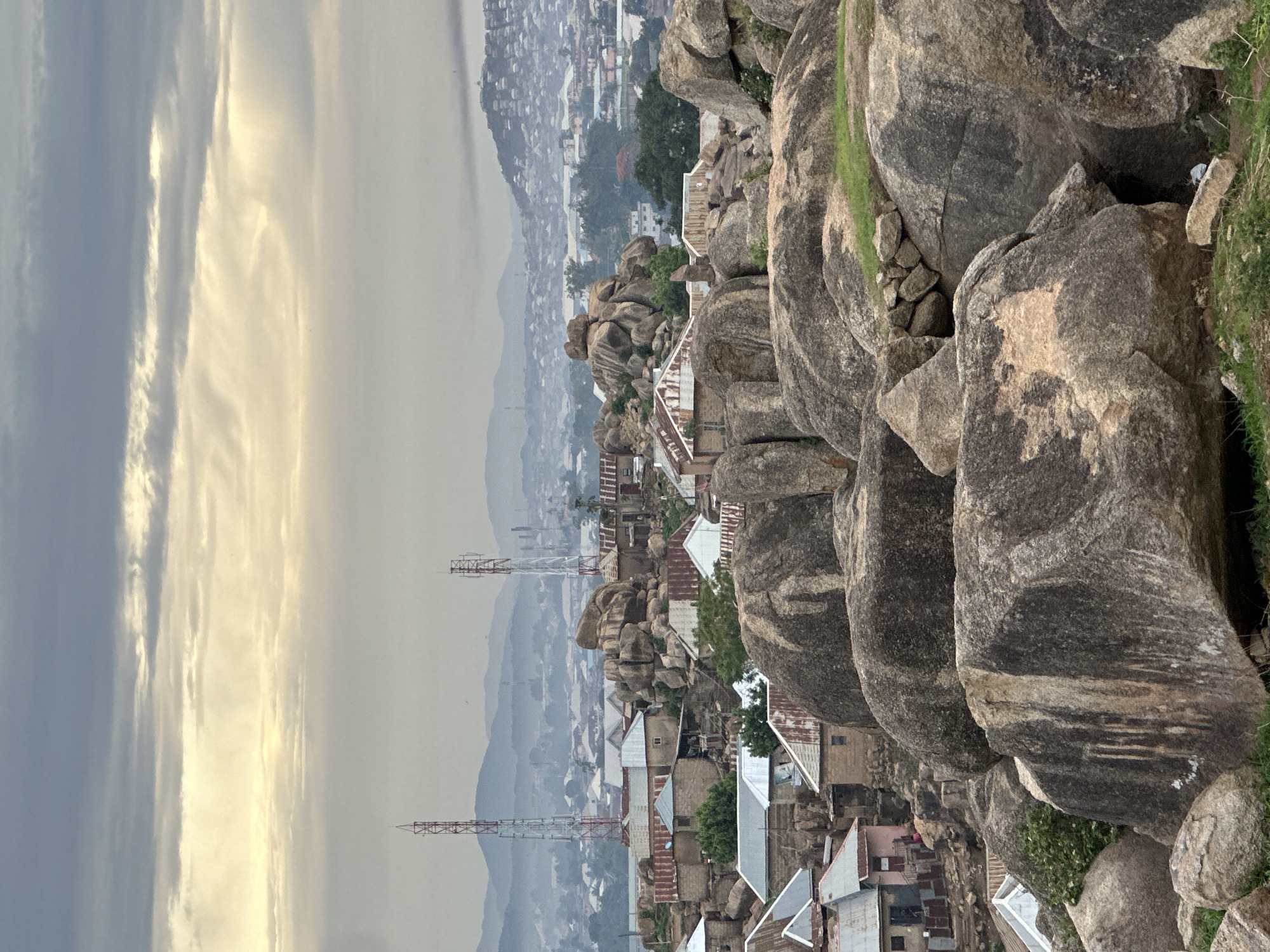 This image presents a panoramic view of the city of Jos, Nigeria, characterized by a rocky landscape under an overcast sky. The horizon features distant, hazy blue-grey mountains. Below the mountains, a sprawling urban area extends into the midground, with numerous structures appearing indistinct due to atmospheric haze.
In the midground, the city becomes clearer, revealing houses with predominantly corrugated metal roofs, exhibiting various states of wear and rust, ranging from silver-grey to reddish-brown. These dwellings are integrated directly into and around massive, weathered granite boulders of varying sizes and shapes. Two prominent red and white lattice-style telecommunication towers stand tall, one on the left and one on the right, rising above the residential structures and rocks. Small patches of green vegetation, including trees and shrubs, are interspersed among the houses and boulders.
The foreground is dominated by large, dark grey to brown granite boulders, some exhibiting streaks of lighter minerals or water erosion marks. Sparse green grass and low-lying vegetation grow in crevices and on flatter surfaces between the rocks. No discernible human figures are present, nor are any specific actions or interactions evident. The overall environment suggests a densely settled area built upon and around a significant geological formation of large rocks, under a sky filled with grey and pale yellow clouds, indicating diffuse or indirect sunlight.