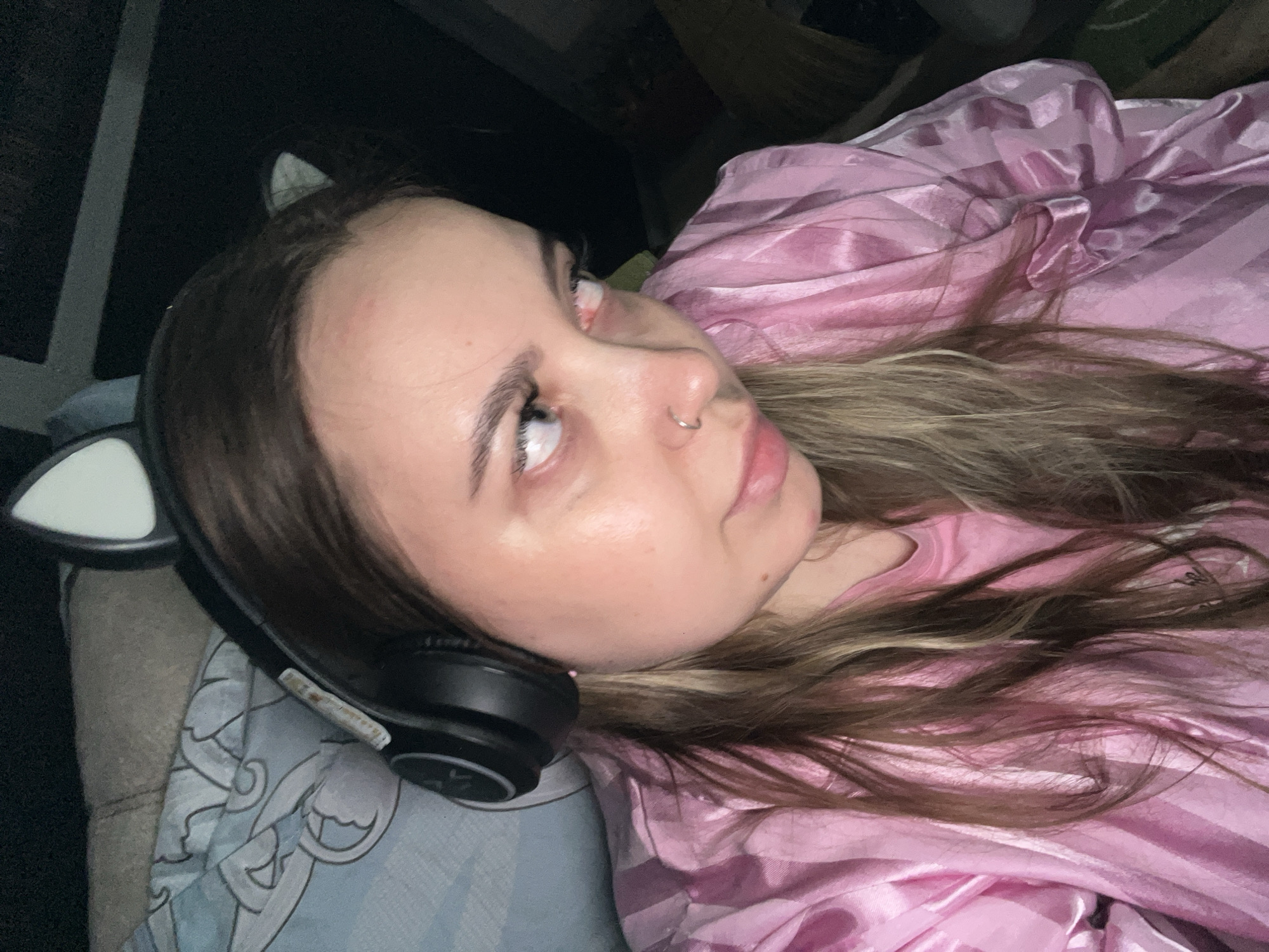 A young woman is lying down, wearing black headphones with cat ear attachments. She has a nose ring and is dressed in a shiny pink top. She is lying on a patterned blue pillow. The location is not discernible from the image.