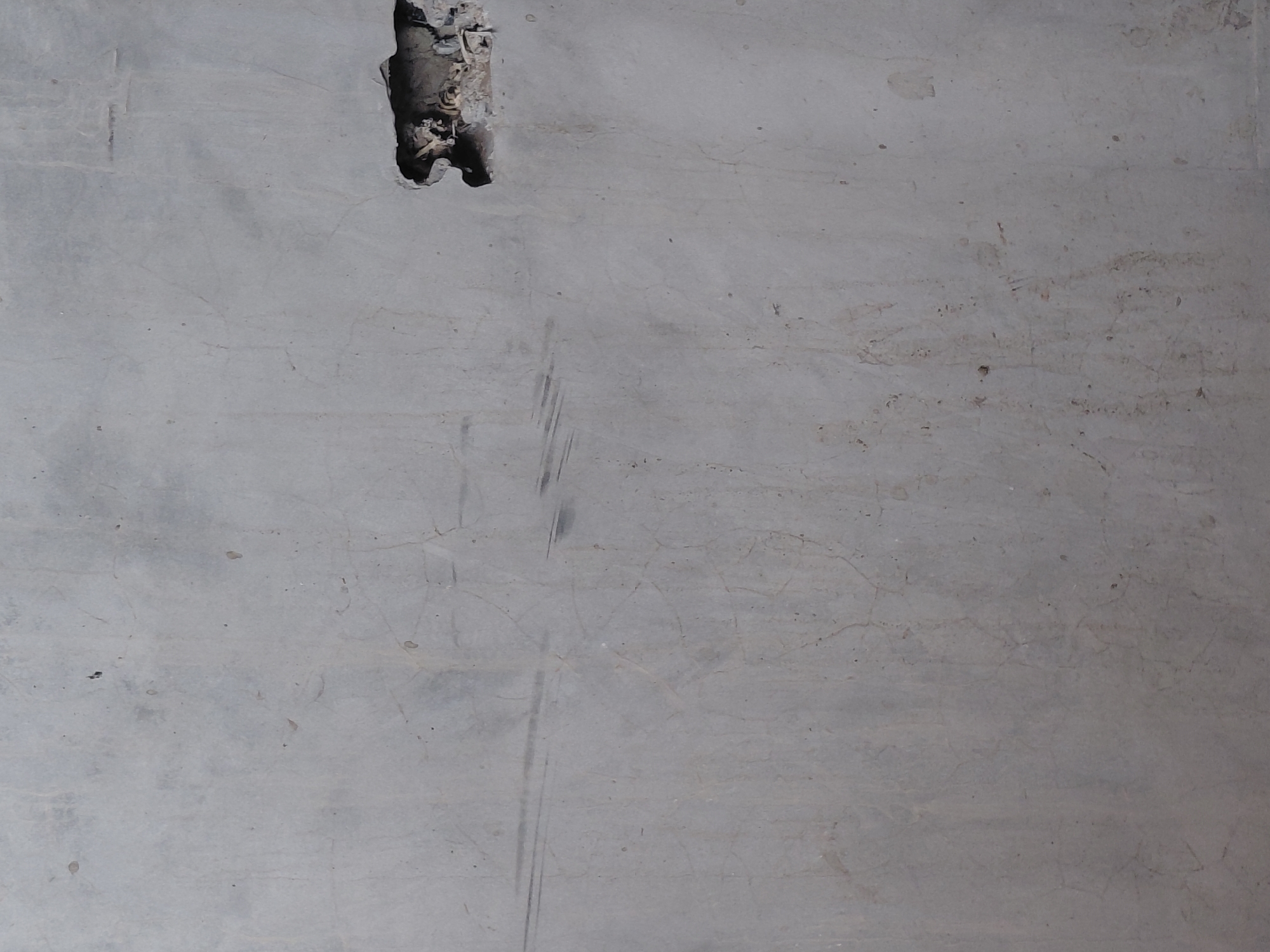 The image shows a textured, light gray concrete wall with minor imperfections. A dark, irregular hole is present in the upper right quadrant of the wall, revealing debris and possibly wiring within. There are several faint scratches and hairline cracks visible across the surface of the concrete, suggesting age and exposure. The lighting is even, indicating an overcast day or an indoor setting with diffused light. No people or specific environmental features of Santa Ana, Venezuela are depicted, the image is a close-up of a damaged wall.