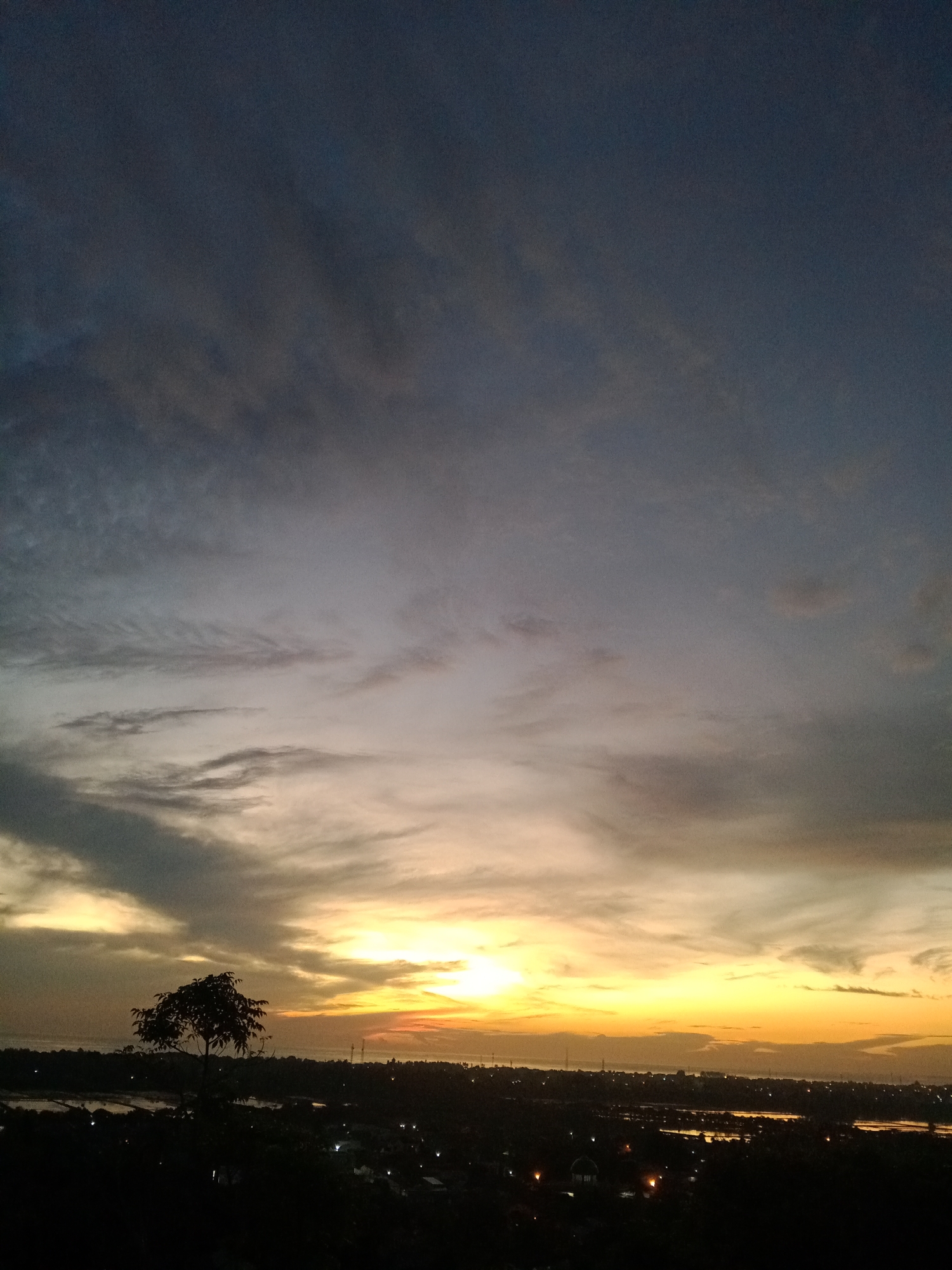 The image captures a sunset scene over the city of Lhokseumawe, Indonesia. The sky exhibits a gradation of colors, from dark blue-gray at the top to yellow and orange near the horizon, indicating the setting sun. Clouds are present, adding texture and complexity to the sky. In the foreground, a silhouette of a tree stands to the left. Below the horizon, there is a dark, obscured view of the city, with some lights visible amidst the darkness. Bodies of water are also visible in the dark landscape. The image gives a sense of a calm, quiet evening in Lhokseumawe.