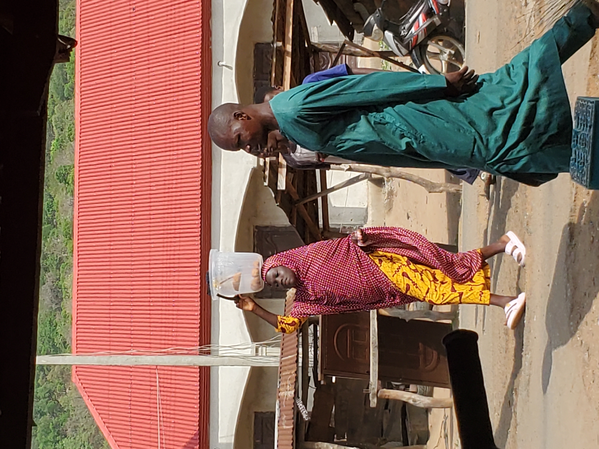 In Mararaba, Nigeria, a woman in a red patterned top, yellow fabric, and white sandals carries a clear plastic container on her head. A man in a green robe stands nearby. A motorcycle is parked in the background. A red corrugated metal roof is visible. The setting appears to be an outdoor area near buildings constructed with wooden and concrete elements.