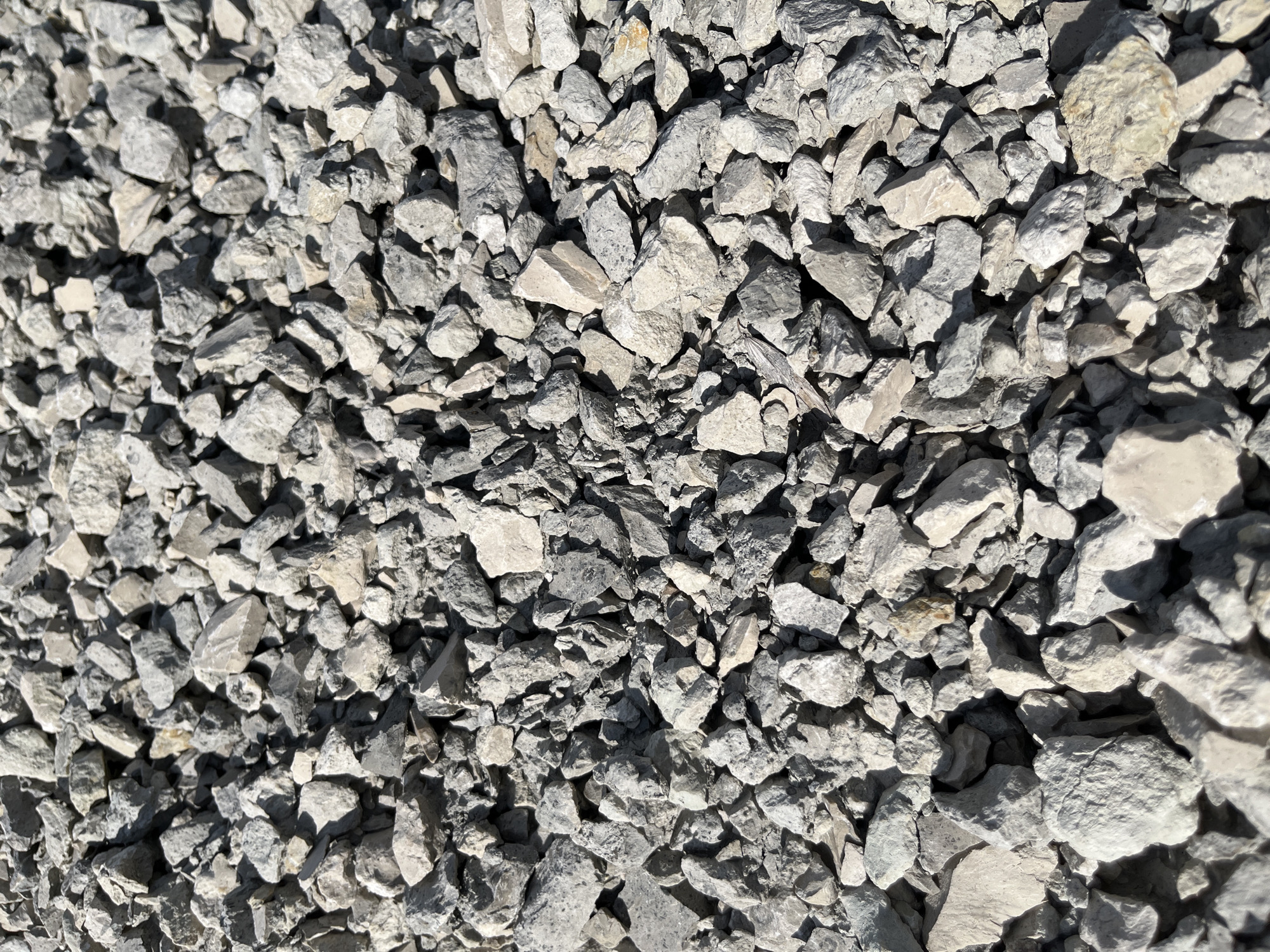 The image is a close-up, overhead view of a pile of crushed rock. The rocks are angular and irregular in shape, with a range of sizes from small gravel to larger chunks. The color palette is predominantly gray and off-white, with some darker gray and occasional lighter, almost yellow-tinged stones. The texture appears rough and coarse. The lighting suggests a sunny day, casting shadows across the surface of the rocks, which adds depth and dimension to the image. The composition is a dense, uniform texture with no discernible patterns or focal points beyond the individual stones themselves. There are no people, animals, or specific landmarks visible, making the location indeterminate. The scene depicts an accumulation of construction aggregate or natural stone fragments.