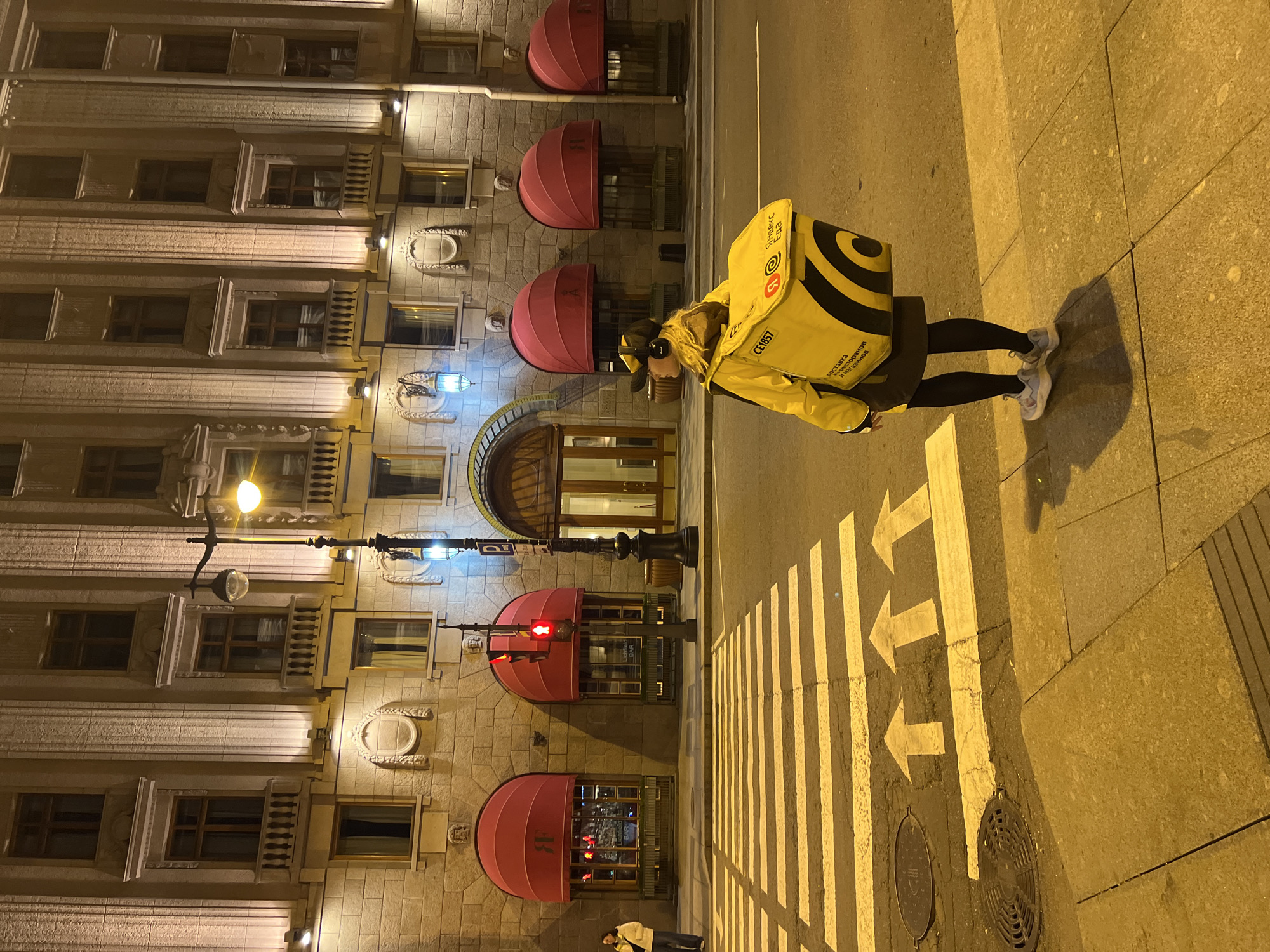 A vertical, nighttime shot depicts a woman in a yellow delivery jacket with a logo-emblazoned yellow backpack walking on a crosswalk in Saint Petersburg, Russia. The woman is seen in profile, facing left, wearing headphones and dark pants with light-colored sneakers. The crosswalk consists of white stripes and directional arrows pointing left. To the left of the crosswalk is a building with multiple windows, illuminated by exterior lights. Pink awnings shade the lower-level windows. A lamppost with a lit light and a red traffic light is situated near the building. The overall scene has a strong yellow-orange cast, typical of nighttime urban lighting.