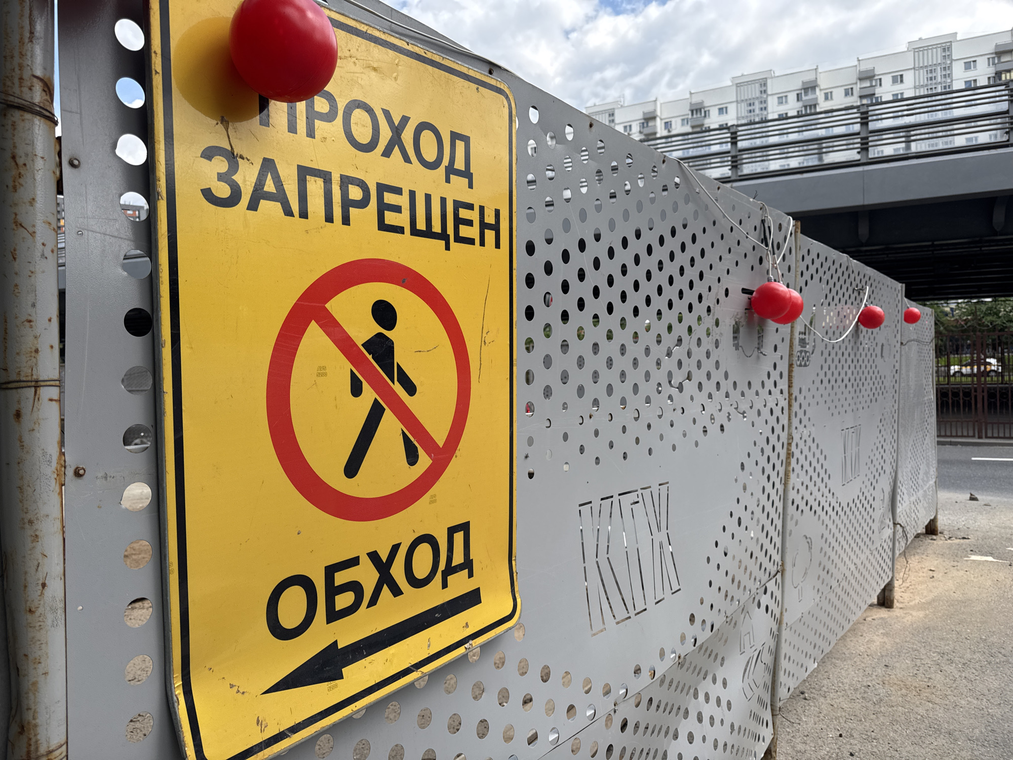 The image shows a temporary construction barrier in the city of Unknown, Unknown. The barrier is made of perforated gray metal and has a yellow sign attached to it. The sign reads "ПРОХОД ЗАПРЕЩЕН" (No Entry) in Russian, along with a symbol of a person crossed out and the word "ОБХОД" (Detour) with a black arrow pointing left. Red warning lights are attached to the barrier with wires. In the background, there is a bridge, a multistory building, and a cloudy sky. The barrier is located on a road, and the overall scene suggests a construction zone.