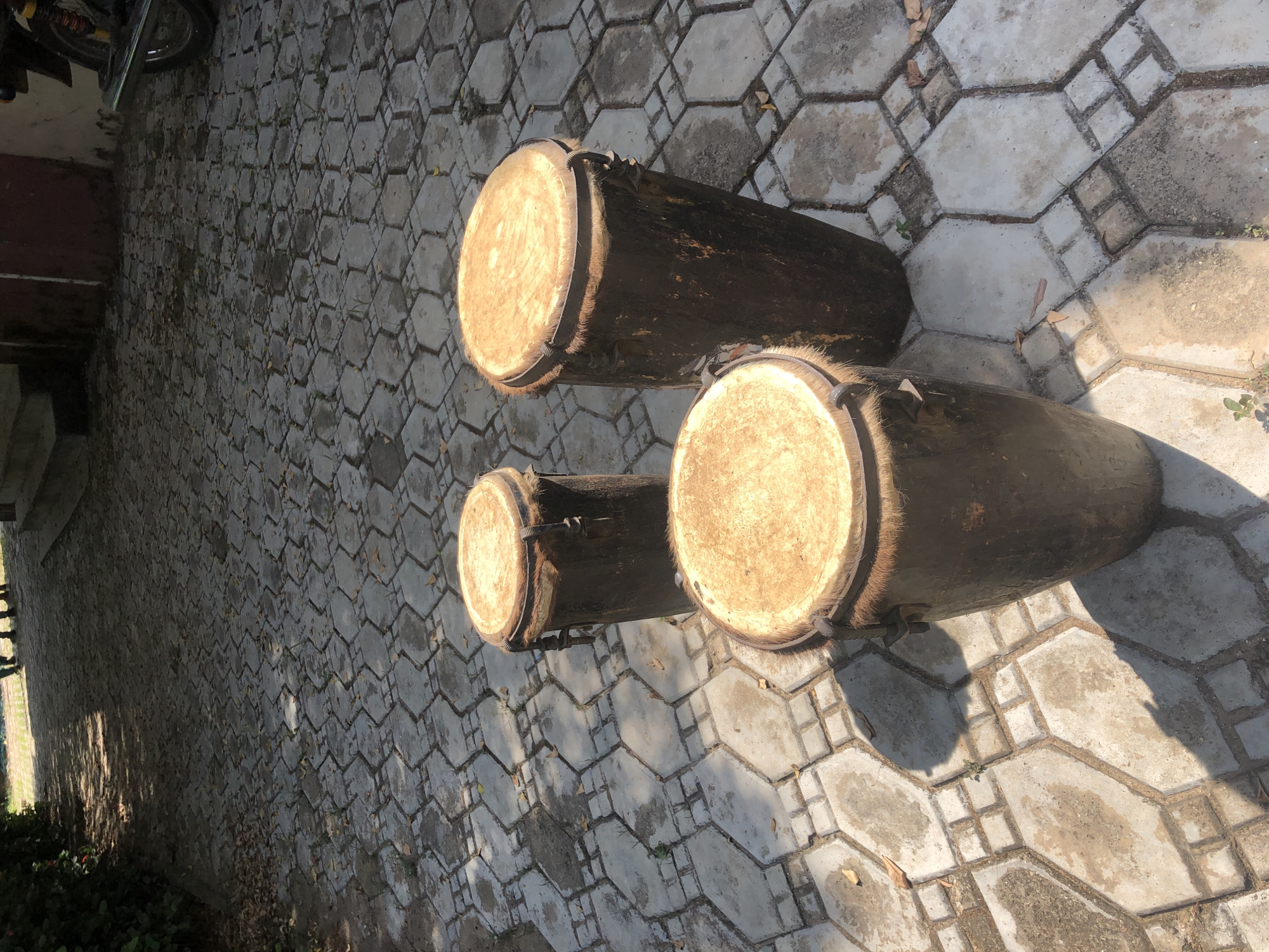 The image shows three traditional African drums lying on a patterned concrete surface. The drums are cylindrical, made of dark wood, with light-colored drumheads stretched across the openings and secured with metal bands. The surface the drums are resting on is composed of interlocking concrete tiles in a diamond-like pattern. The location of the image is Akwanga, Nigeria. A partially visible motorcycle is in the background.