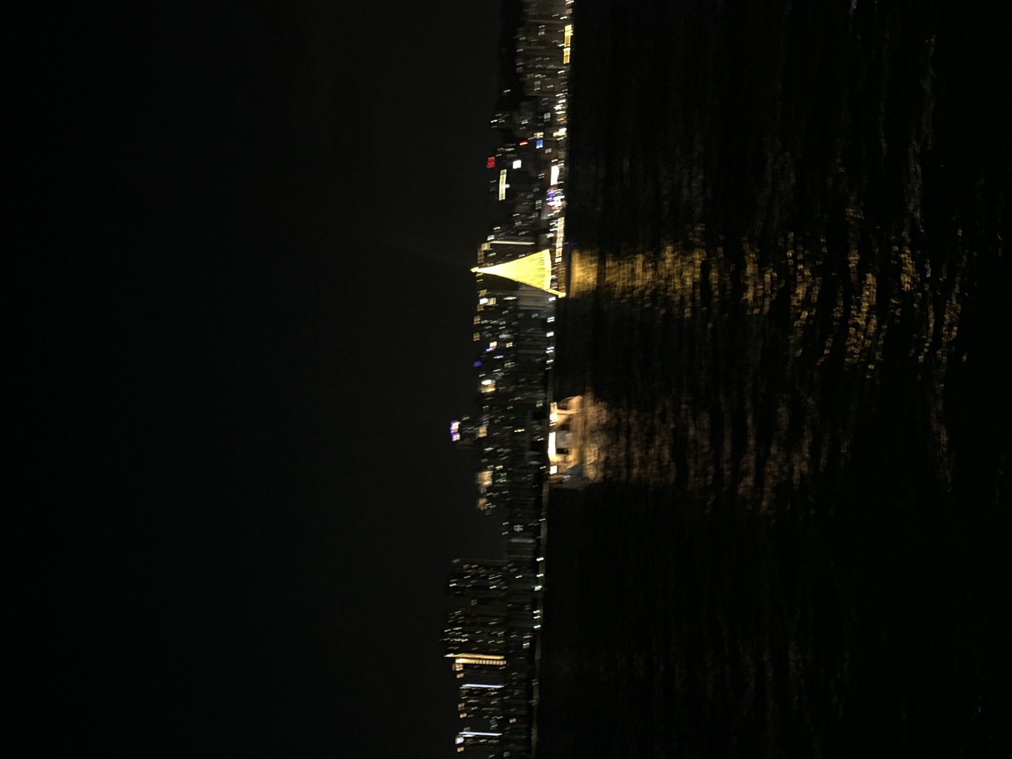 An unfocused nighttime view of a waterfront cityscape in Busan, South Korea. A body of water occupies the lower half of the image, reflecting scattered golden lights from the buildings and a festive, tree-shaped illuminated structure in the middle ground. The background features a dense line of illuminated high-rise buildings, indicative of a major urban center. A boat with lights is visible on the water, to the left of the illuminated tree. The overall impression is one of a vibrant city at night, viewed from a distance across the water.