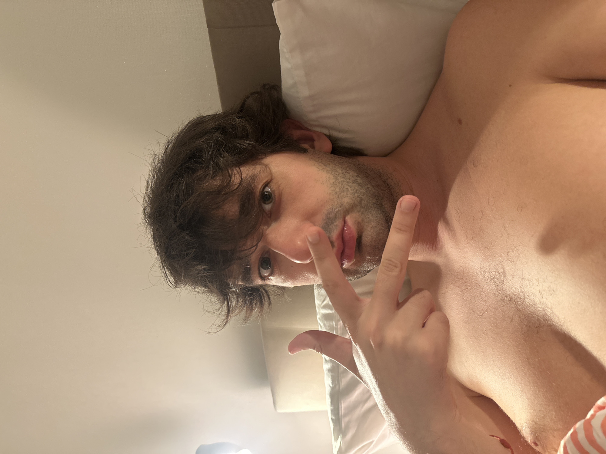 The image shows a close-up, selfie-style shot of a man lying in bed. He has dark, messy hair and is looking directly at the camera with his eyes open. His left hand is raised to his face, with his index and middle fingers extended in a "V" shape, positioned near his nose. He is shirtless and appears to be in an indoor setting, likely a bedroom, with a white pillow and a brown headboard visible in the background. The lighting suggests it is nighttime or a dimly lit room, possibly with a lamp on. There is no identifiable information that places the image in Moscow, Russia.