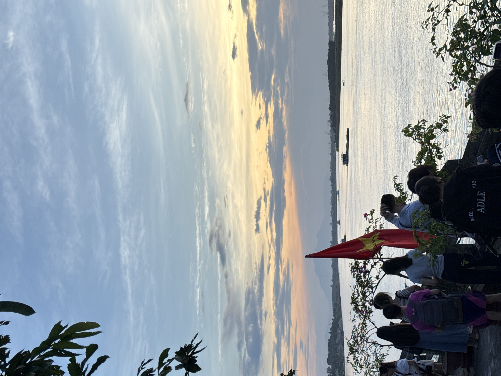 At dusk, the image shows a group of people with a flag on the bank of the Red River (Sông Hồng) in Hà Nội, Vietnam. The sky is a gradient of blue to orange, and clouds reflect the setting sun. The river is wide and calm. In the middle distance, boats are visible. The flag appears to be the flag of Vietnam. The people are standing near flowering bushes, taking pictures or looking out at the view.