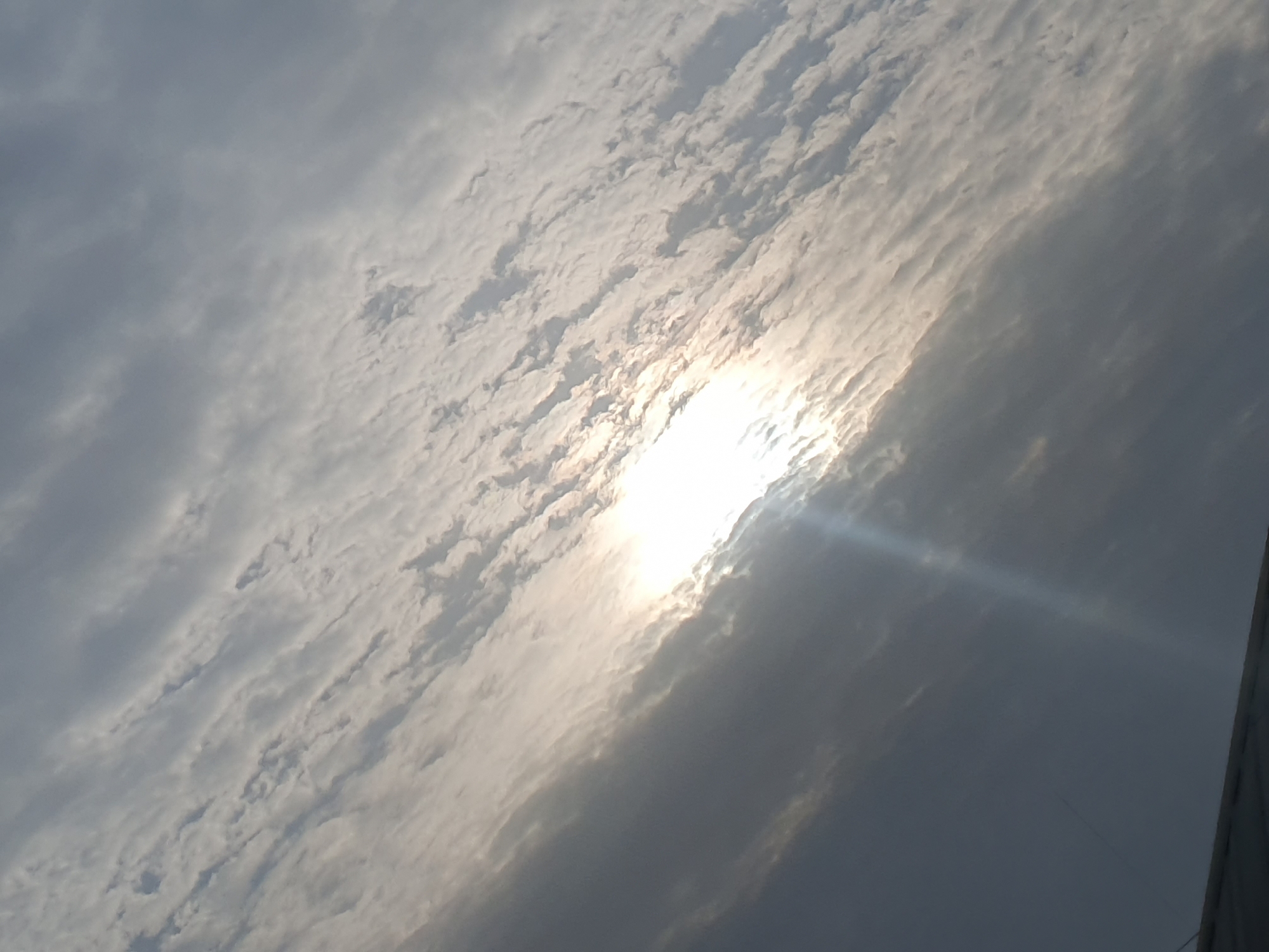 The image shows a view of the sky with clouds and the sun peeking through. The sun is bright and appears to be low in the sky, casting a warm glow. The clouds are a mix of white and gray, with some areas appearing thicker and more opaque than others. The scene is tilted, suggesting the photo was taken from an angle. In the lower portion of the image, a dark, flat surface is visible, possibly a rooftop or a structure. The overall impression is one of a dramatic sky with the sun breaking through the clouds. The location is specified as Gura Top, Nigeria.