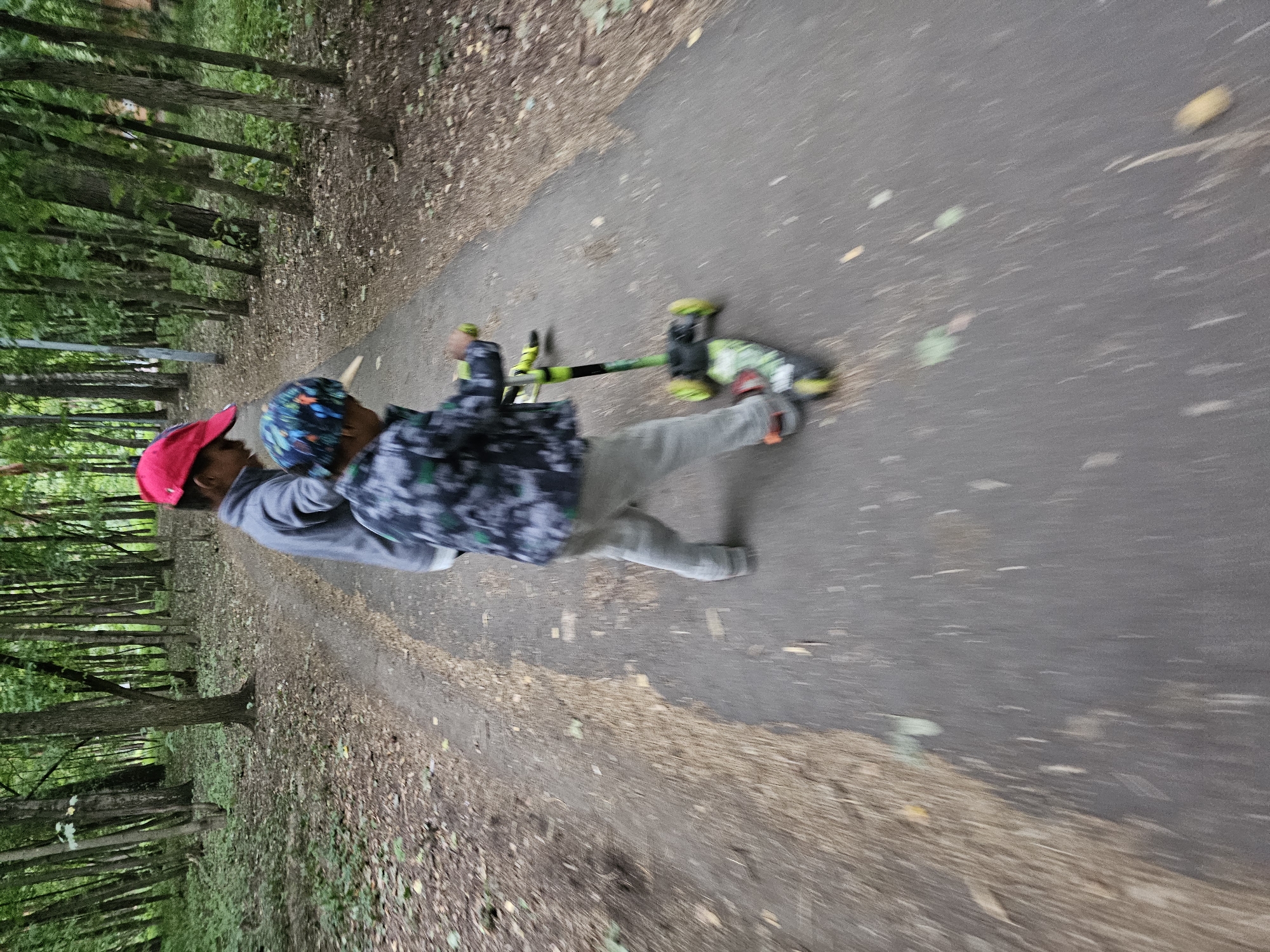 Two children are in a park in Khimki, Russia. The child in the foreground is riding a green and black three-wheeled scooter. They are wearing a camouflage jacket, grey pants, and a patterned cap. The child behind them is wearing a grey jacket and a red baseball cap. The children are on a paved path surrounded by trees. The ground is covered with fallen leaves. The image is slightly blurred, suggesting movement.