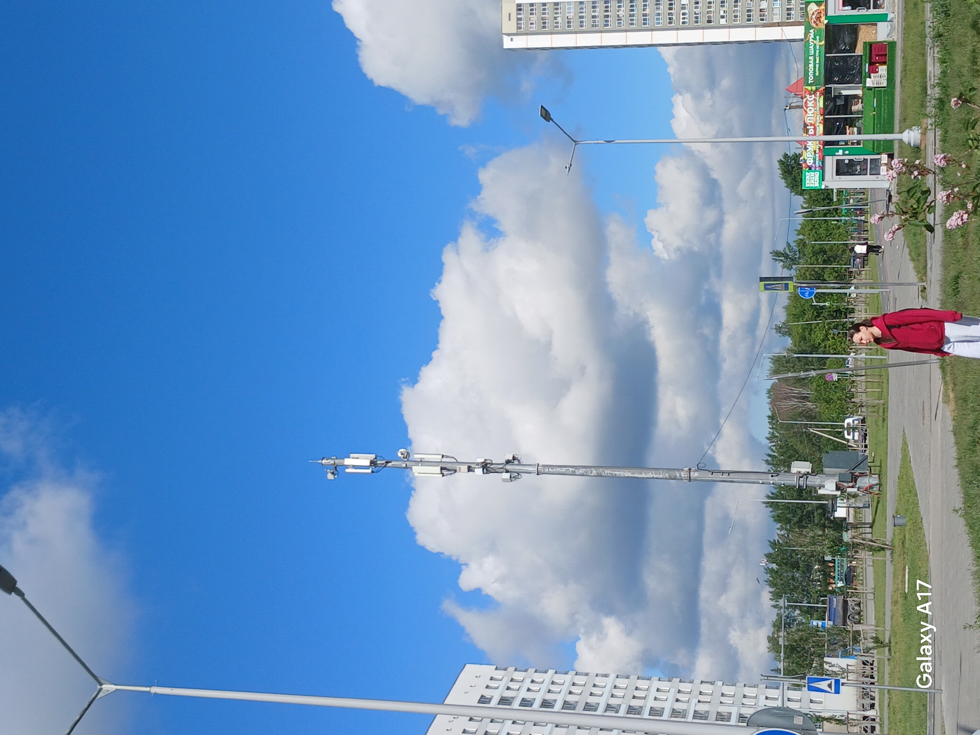 The image captures a vibrant urban street scene on a bright, cloudy day, likely in Yekaterinburg, Russia, based on the provided context and visible Cyrillic text. The sky dominates the left side of the frame, a deep blue adorned with large, fluffy cumulus clouds.
In the mid-ground, a tall pole equipped with multiple antennas, resembling a cell tower or communication mast, stands prominently. Several street lights are also visible. To the right, a woman wearing a red long-sleeved top and white pants stands on a paved sidewalk, looking towards the camera.
The street level features a paved road bordered by green grass and some flowering bushes with small pink blossoms. Along the street, a cluster of brightly colored commercial kiosks or small shops is visible. One kiosk is primarily green and black, displaying text "ТОРГОВАЯ ШАУРМА" (Commercial Shawarma), "СУШИ РОЛЛЫ" (Sushi Rolls), and "ФРИ ДРИНКС" (Fries Drinks or Free Drinks). Another sign shows "БЛЮКС" (BLUX).
In the background, two multi-story residential buildings with numerous windows rise, one on the upper right and another partially visible on the lower left, indicating a densely populated urban area. Trees line the street in the distance, and various street signs, including a blue circular sign and a yellow diamond-shaped sign, are present. Small glimpses of vehicles can be seen further down the road. The time of day appears to be daytime, given the clear, well-lit conditions. A watermark "Galaxy A17" is visible in the bottom right corner, indicating the device used to capture the image.