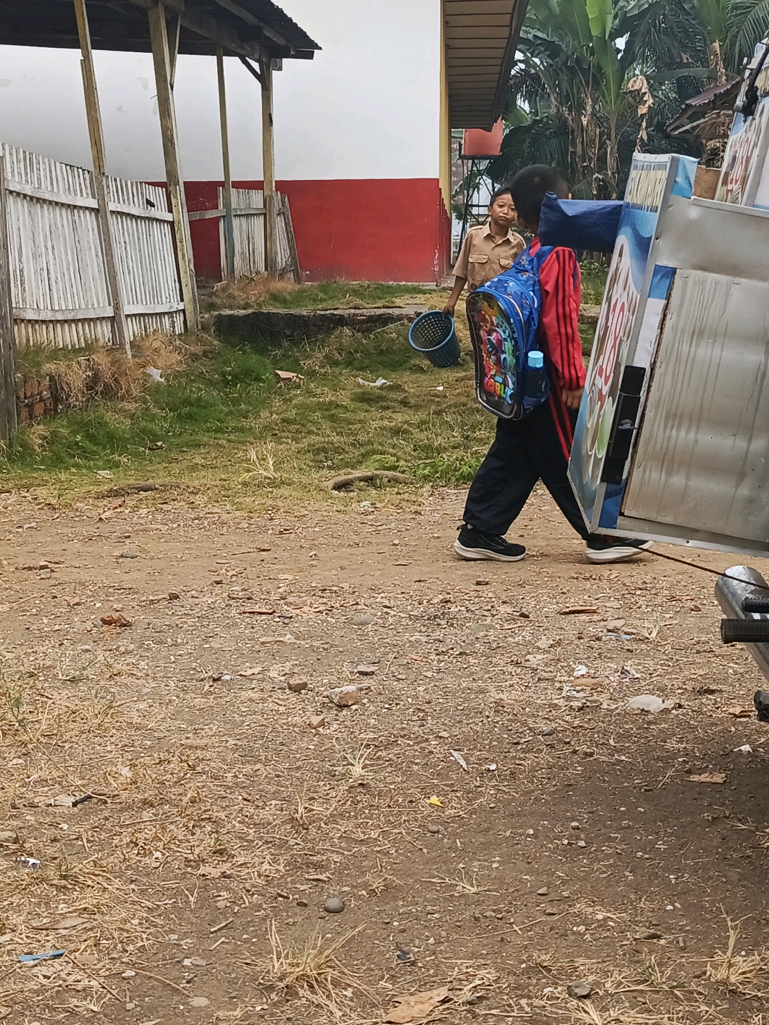 In Megang Sakti II, Indonesia, two young boys are visible on a dirt path near a building with a red wall and a covered walkway. One boy, wearing a tan uniform and holding a blue basket, stands on the left, looking towards the right. To his right, another boy, wearing a red jacket with stripes, black pants, and carrying a colorful backpack and a blue water bottle, walks away from the camera, his back partially turned. He is near large, metallic panels, one of which has a blue and white graphic. The environment is unkempt, with dry grass, scattered debris, and gravel on the ground.