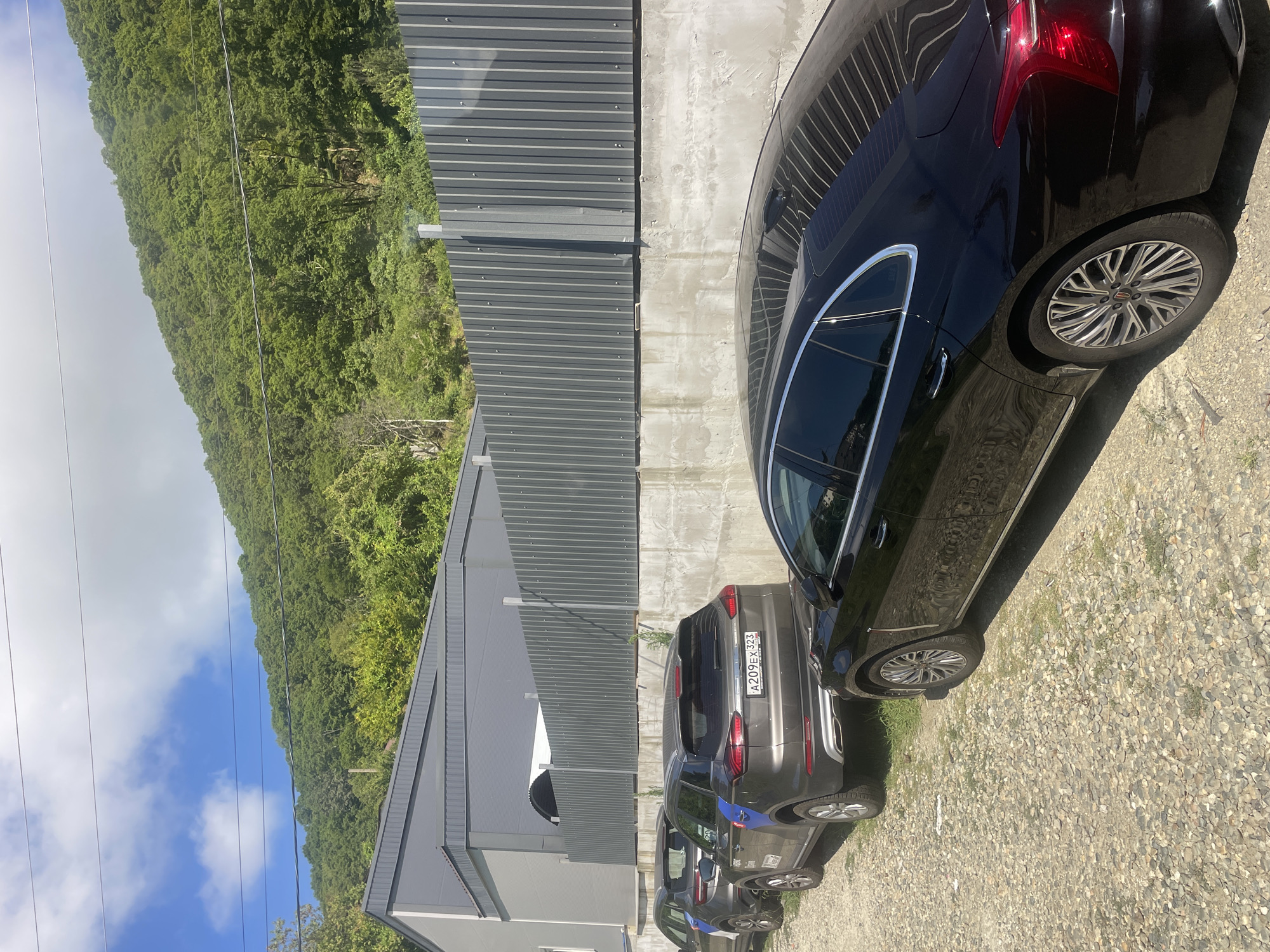 The image shows a row of parked cars on a gravel lot in Baranovka, Russia. The cars are parked next to a concrete wall topped with a corrugated metal fence. Behind the fence, a modern gray building is visible, and further back, a lush green forested hillside rises steeply under a bright blue sky with scattered clouds. The cars appear to be in good condition and are parked in a line, suggesting a parking area. The overall scene depicts a sunny day in a semi-urban or semi-rural setting with a focus on the parked vehicles and the natural landscape.