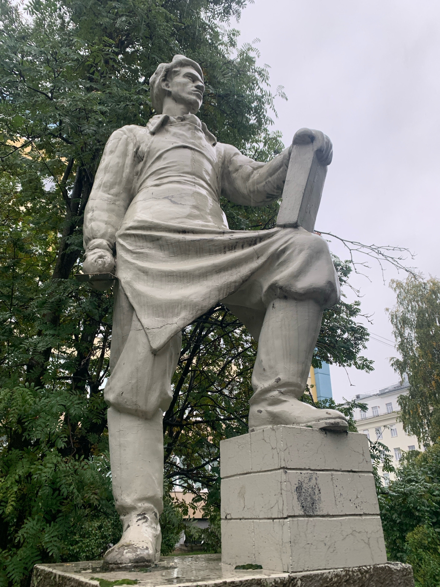A full-length, light-colored concrete statue of a man stands in Arkhangelsk, Russia. The man is depicted in work clothes, with a tool or object held in his right hand and his left leg resting on a block. The statue is situated outdoors, with lush green trees in the background and a cloudy sky overhead. Buildings can be seen in the distance. The statue's surface appears to be weathered, with some cracking and discoloration visible. The man's posture and attire suggest he is a laborer or craftsman.