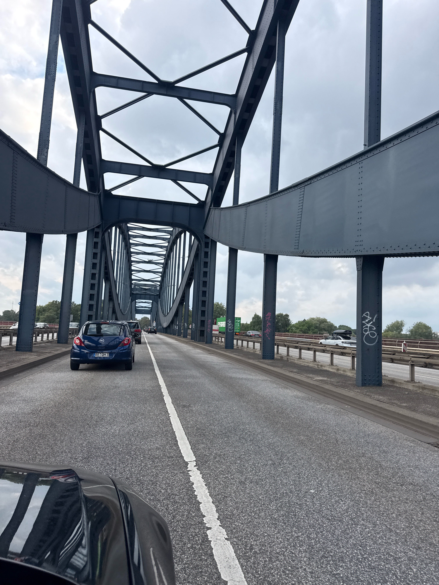 The image shows a view from a car driving on a multi-lane bridge in Hamburg, Germany, likely over water, under a cloudy sky. The bridge is constructed with dark grey steel girders forming an arching structure overhead, with visible rivets and supporting pillars. Several cars are visible on the bridge, including a blue Opel Corsa in the foreground. The road surface is asphalt with white lane markings. In the background, trees and some buildings can be seen on the banks. The overall impression is one of urban infrastructure and transportation.