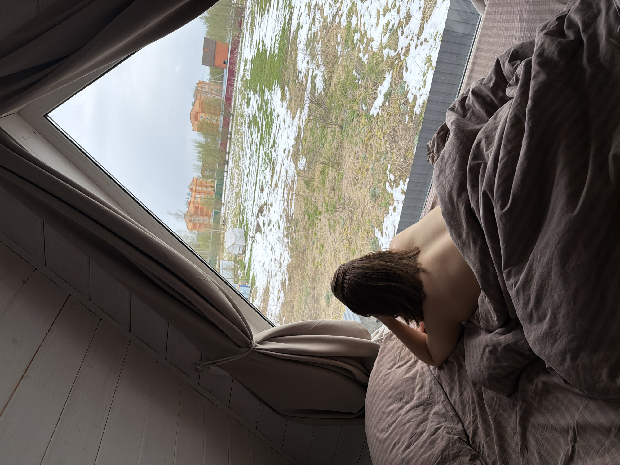 An intimate indoor scene depicts a person lying in bed and looking out a large, angled window in Бунятино, Russia. The person, who has short brown hair and a bare back, is partially covered by a rumpled, dark grey or brown duvet. They are positioned on their stomach, resting their head on one arm, gazing out at the view.
The room features a bed with striped bedding matching the duvet's color. To the left, a white, slatted wooden wall suggests an attic or a room with an angled ceiling. Light grey curtains are drawn back and tied on either side of the window frame.
Through the window, an outdoor landscape unfolds under an overcast sky. The ground is covered in patchy snow, interspersed with dry brown grass and some areas of early green growth, indicative of late winter or early spring. In the mid-ground, a few bare trees and shrubs are visible. Further in the distance, several multi-story brick buildings, likely apartment blocks, stand against the horizon, surrounded by sparse vegetation. The scene evokes a quiet, contemplative moment, possibly in the morning, observing the cold, transitional weather outside. No text is visible in the image.