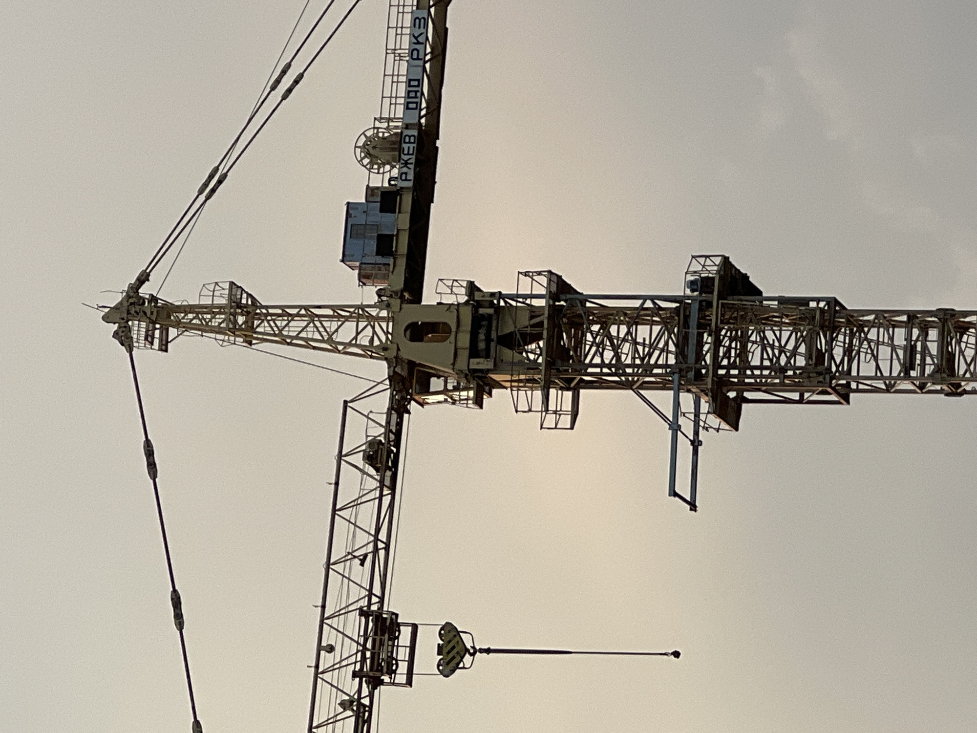 This is an outdoor, low-angle shot of a large construction crane against a bright, overcast sky. The image is oriented vertically, with the top of the crane appearing at the bottom of the frame and vice versa. The crane's structure is complex, made of metal lattice work. A key component, the operator's cab, is visible, as are various wires, pulleys, and a long jib arm extending horizontally. The light suggests it is daytime, possibly late afternoon or early morning, given the soft, diffused light. There are no people visible. The scene is clearly a construction site, as indicated by the presence of the crane. Some text is visible on a vertical section of the crane's tower; it appears to be in Cyrillic script and reads "РЖЕВ 090 PK3". The overcast sky suggests potentially cloudy or hazy weather. The overall impression is one of industrial activity and large-scale engineering.