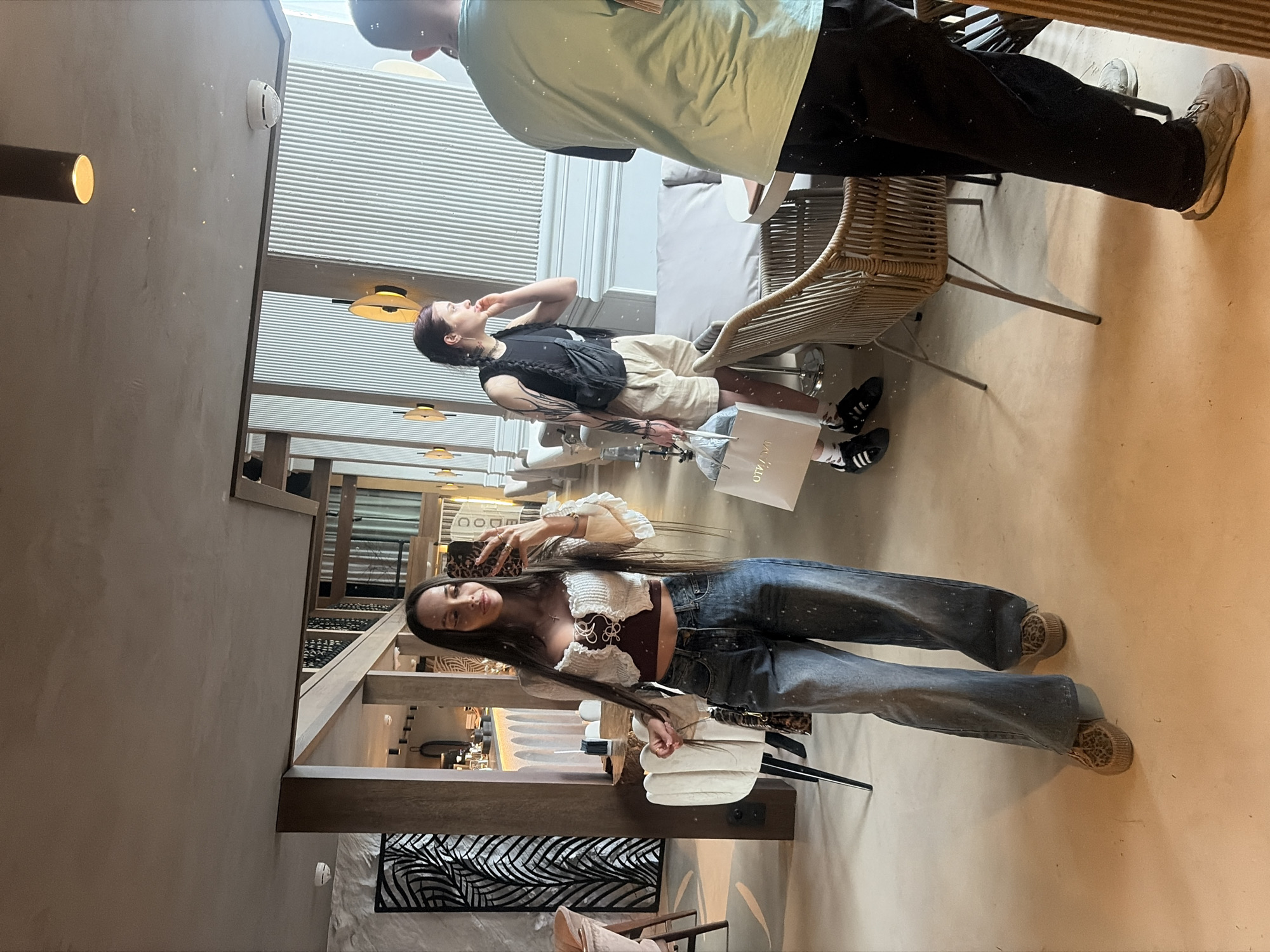 In the city of Saint Petersburg, Russia, a woman stands in what appears to be a modern cafe or restaurant, taking a mirror selfie. She has long dark hair and is wearing a white ruffled crop top over a maroon bralette, paired with wide-leg denim jeans. Her feet are clad in leopard-print platform shoes. To her right, another woman with dark hair braided into two pigtails is seen in the reflection, wearing a black tank top, beige shorts, and black sneakers. She is holding a white shopping bag. A third person, a man, is partially visible in the far right of the reflection, wearing a light blue t-shirt and dark pants. The interior features contemporary decor, including textured walls, minimalist furniture, and ambient lighting. The overall scene suggests a casual outing or shopping trip.