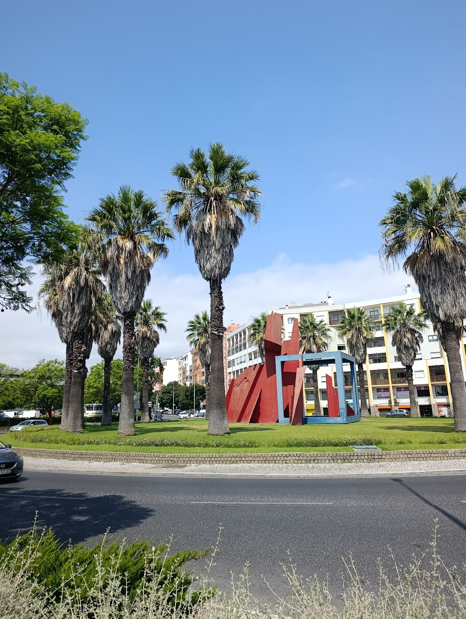 This is a medium-angle, eye-level, outdoor photograph taken on a sunny day in Setúbal, Portugal. The scene captures a grassy circular median with several palm trees and a modern sculpture. The sculpture is composed of bright red angular shapes and a blue rectangular frame. Behind the median, there are buildings with cream-colored facades and colorful accents, typical of urban architecture. A street with a few cars and a white bus is visible in the background, indicating a city environment. In the foreground, a dark gray asphalt road runs horizontally across the bottom of the frame, with a paved pedestrian walkway and low stone wall bordering the median. Green shrubs are in the lower right corner. The sky is clear and blue with a few wispy clouds. The photograph is likely taken from a moving vehicle due to the slight blur and perspective. The overall impression is a vibrant urban landscape with a tropical touch.