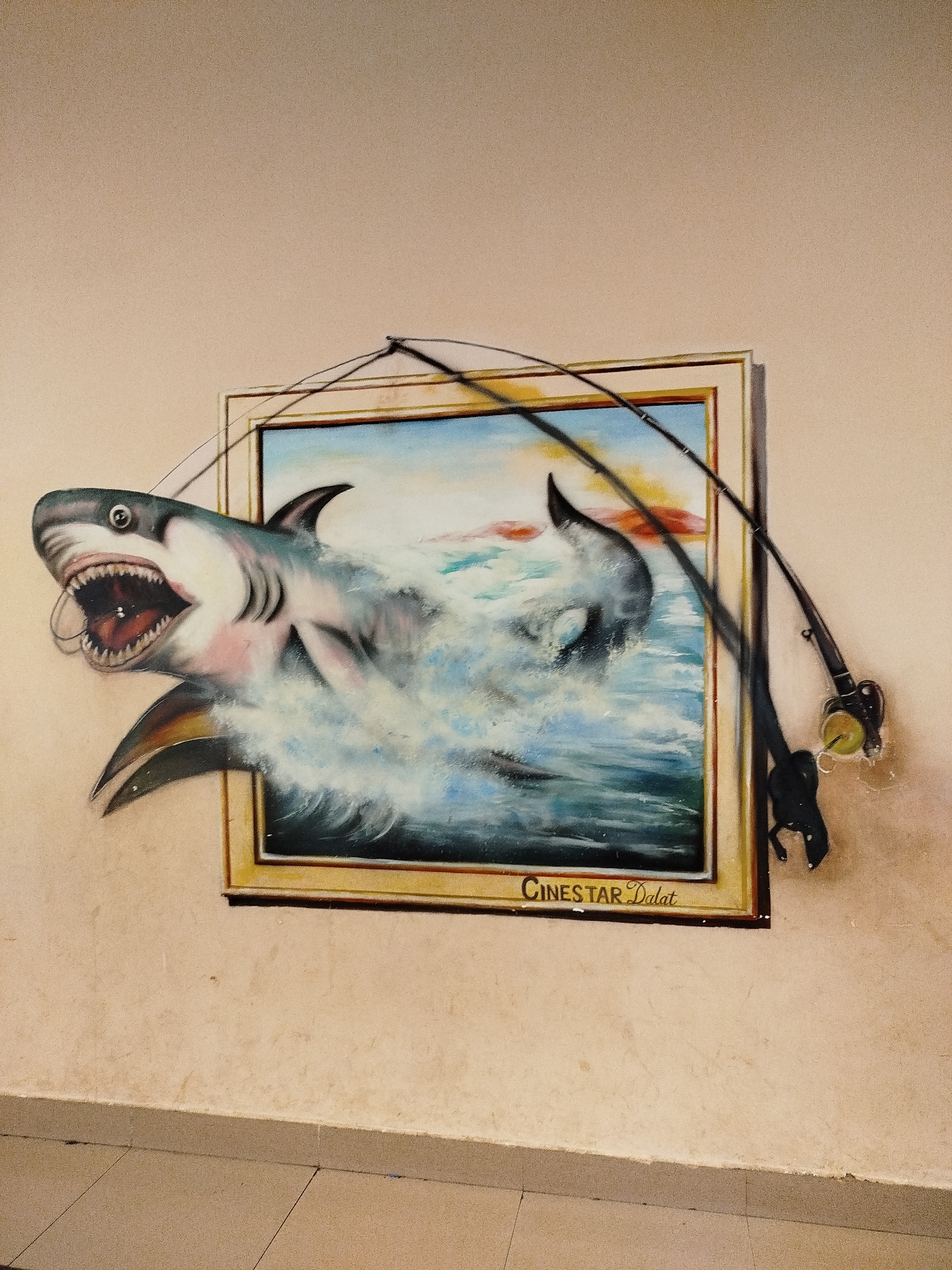 A low-angle shot captures a trompe-l'oeil painting on a textured beige wall. The painting is of a shark leaping out of the sea, as though escaping the confines of the gold-framed artwork. A fishing rod is visually integrated into the scene, with part of the rod and reel extending outside the frame, enhancing the illusion of depth. The words "CINESTAR Dalat" are painted at the bottom of the frame. The lower part of the image shows a tiled floor and wall baseboard. The location of this artwork is Unknown, Unknown.