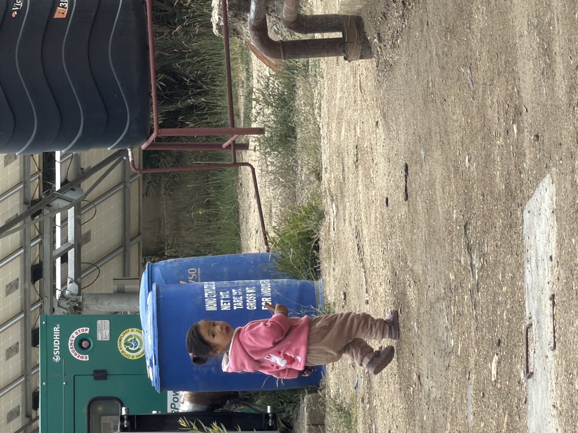 A young girl in a pink hooded sweatshirt and tan pants walks away from the camera on a dirt path. Behind her, a large blue cylindrical tank is visible, with text printed on its side. To the left, a green generator with "SUDHIR" written on it and a red "EMERGENCY STOP" button is present. In the background, solar panels are mounted on a metal frame, and a large black water tank sits on a red metal stand. The environment appears to be an outdoor industrial or storage area, possibly near a settlement, with sparse vegetation and a dusty ground. The overall scene suggests a rural or semi-industrial setting.