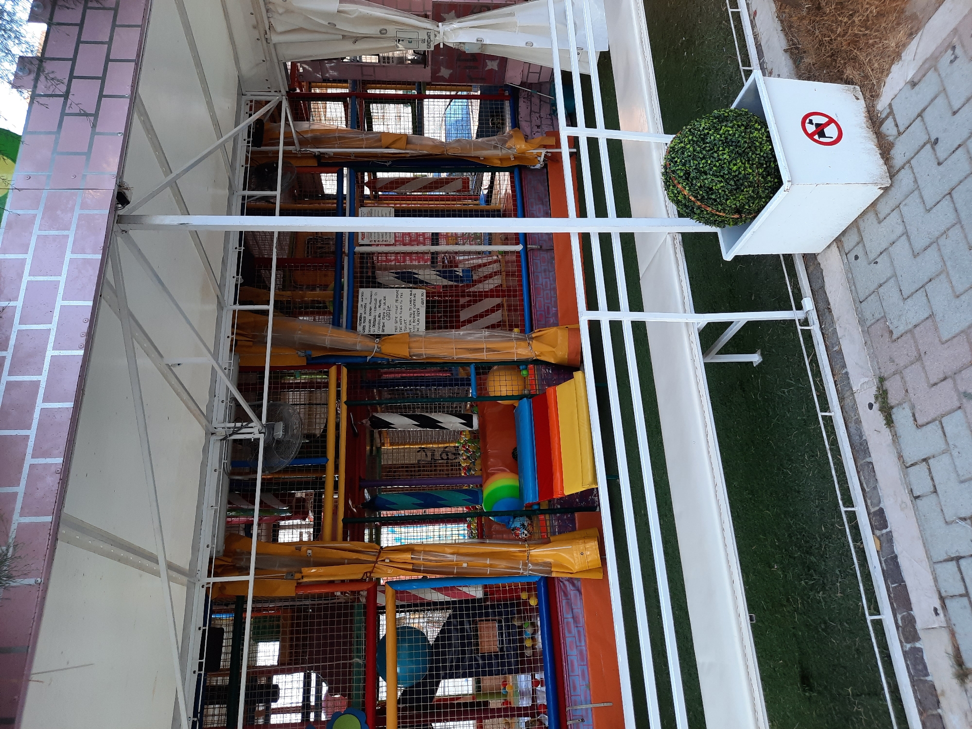 The image shows an outdoor children's play area in Pesaro, Italy. The play structure is a multi-level soft play area with slides, tunnels, and ball pits, enclosed by netting and brightly colored padded surfaces. Yellow curtains hang from the structure. In the foreground, a white bench runs along a white railing. To the right of the bench, a white planter contains a spherical green artificial plant and has a red and white sign indicating "no dogs". The play area is covered by a white awning supported by metal poles. The background shows some trees and buildings. The overall scene suggests a family-friendly outdoor entertainment venue.