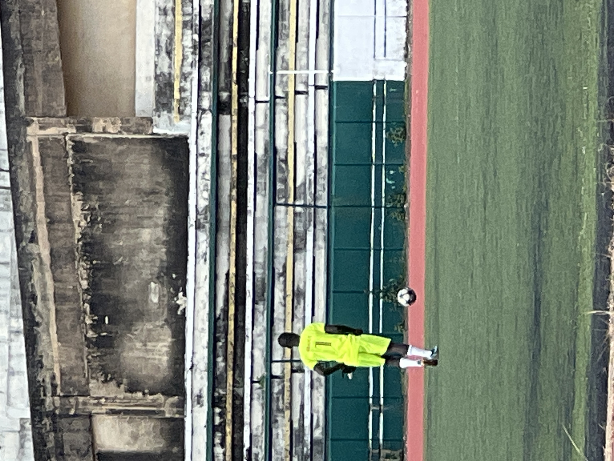 In New Stadium, Nigeria, a lone male soccer player wearing a neon yellow jersey with the number '1' on the back and matching shorts stands on a green artificial turf field. He is facing away from the camera, his left hand on his hip, and his right foot is positioned near a black and white soccer ball. In the background, there are weathered bleachers with green and yellow stripes, and the concrete structure of a stadium with evidence of wear and tear. A red track runs along the edge of the field.