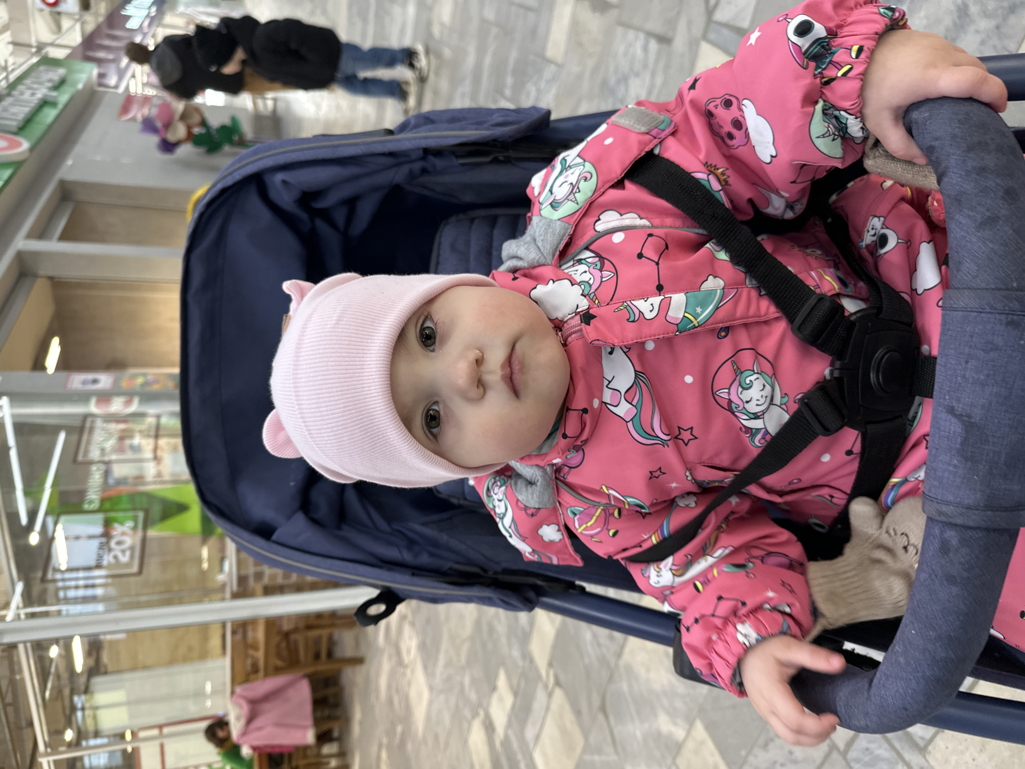 A baby wearing a pink hat with ears and a pink jacket with unicorn patterns sits in a blue stroller. The stroller is indoors on a tiled floor, possibly in a shopping mall or store in Chaikovsky, Russia. People are seen in the background near a store or display with green accents.