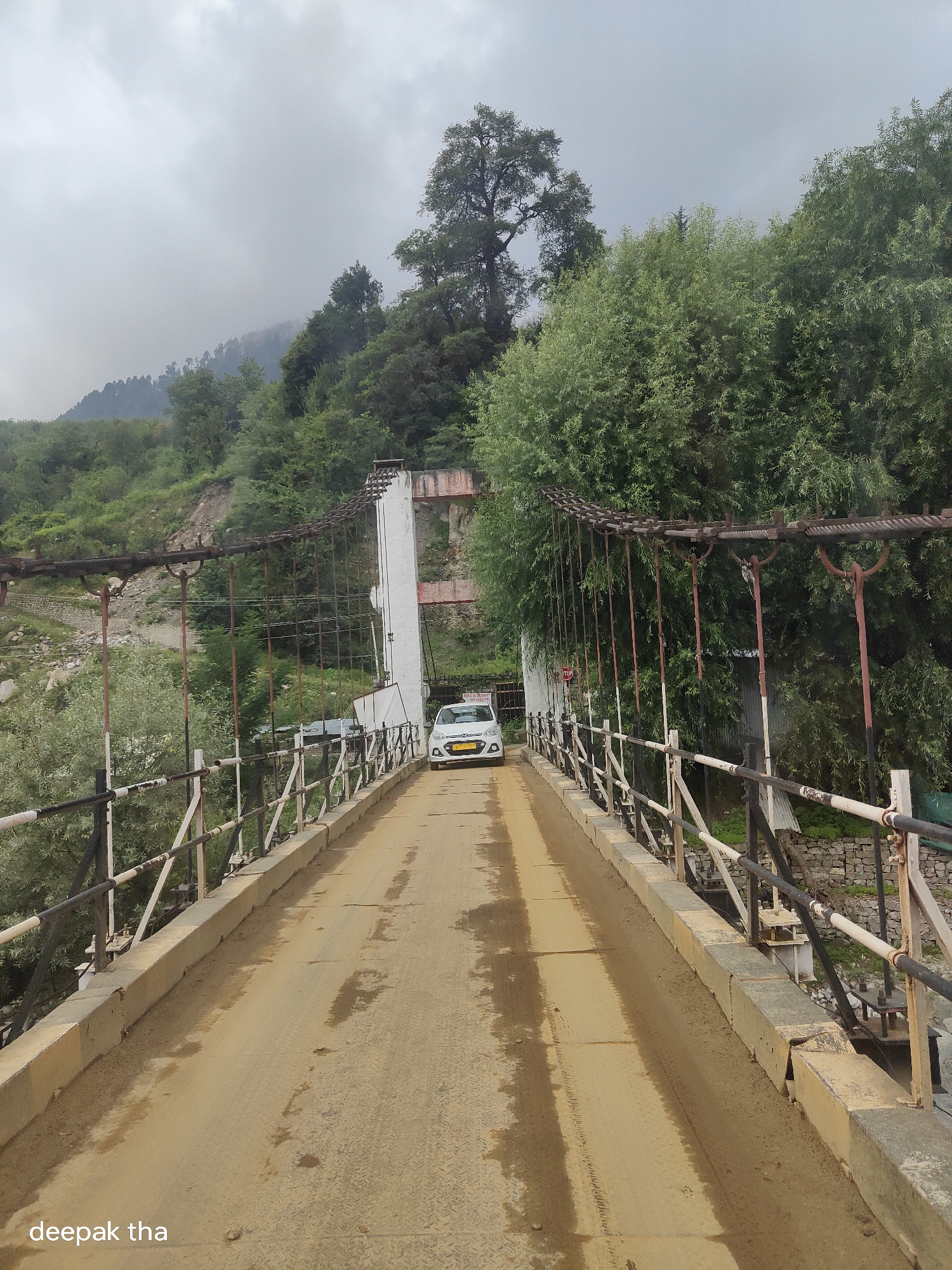 This is a vertical, medium shot taken from a bridge looking forward towards a white car. The bridge, which appears to be constructed of metal and concrete, has a light brown, sandy surface and is lined with a black and white railing. A white sedan is driving across the bridge towards the viewer, its headlights illuminated. The bridge is supported by large white pillars and thick cables that stretch across the frame. In the background, lush green trees and a mountainous landscape are visible under a cloudy sky. The lower left corner of the image contains the text "deepak tha" in white lettering. The scene appears to be in a natural, possibly rural setting.