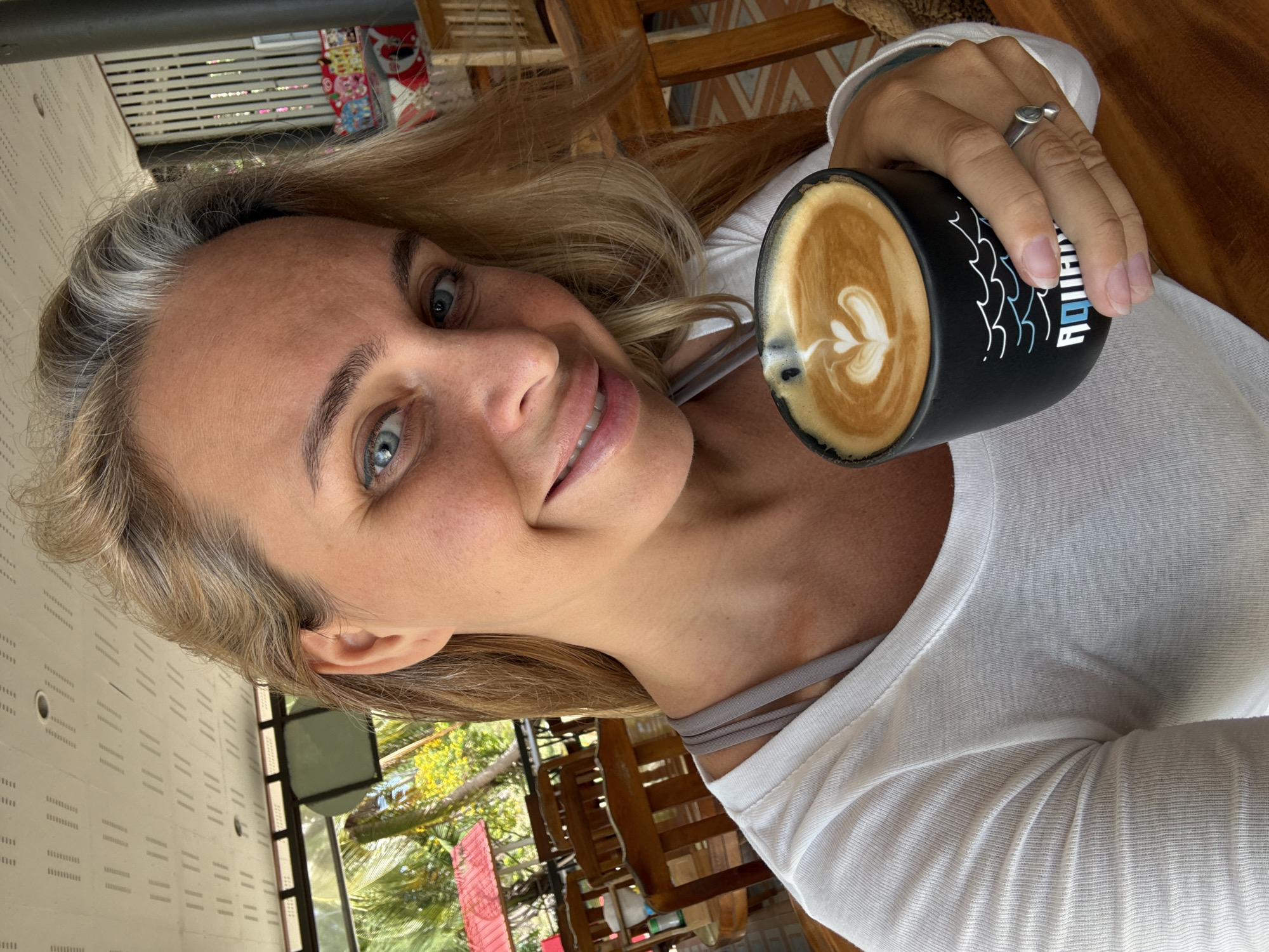 A blonde woman with blue eyes smiles into the camera, holding a black coffee mug with latte art in her right hand. She wears a white t-shirt and a grey bra. The background shows a wooden table and chairs, suggesting an outdoor cafe setting. Palm trees and buildings are visible in the distance. The ceiling above is made of white panels with a repetitive pattern. The woman appears to be at an outdoor cafe on the island of Koh Samui in Thailand.
