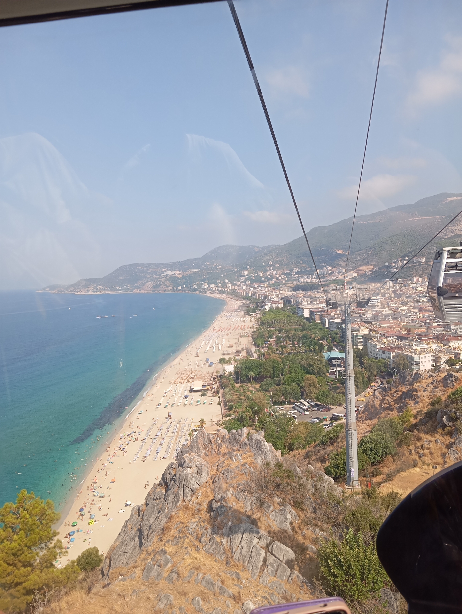 The image is a high-angle shot from a cable car overlooking a coastal city. The foreground shows a rocky, dry hillside with sparse vegetation and the edge of the cable car cabin. Below, a long, sandy beach is crowded with beachgoers, lined with rows of umbrellas and lounge chairs. The turquoise ocean stretches out to the horizon, with several boats visible on the water. Beyond the beach, the city is densely populated with buildings, some with red-tiled roofs, cascading up the hillsides towards the mountains in the background. A tall, grey support tower for the cable car runs vertically through the scene, with cables extending upwards. The sky is a clear blue with a few wispy clouds. The overall scene depicts a popular tourist destination on a sunny day.