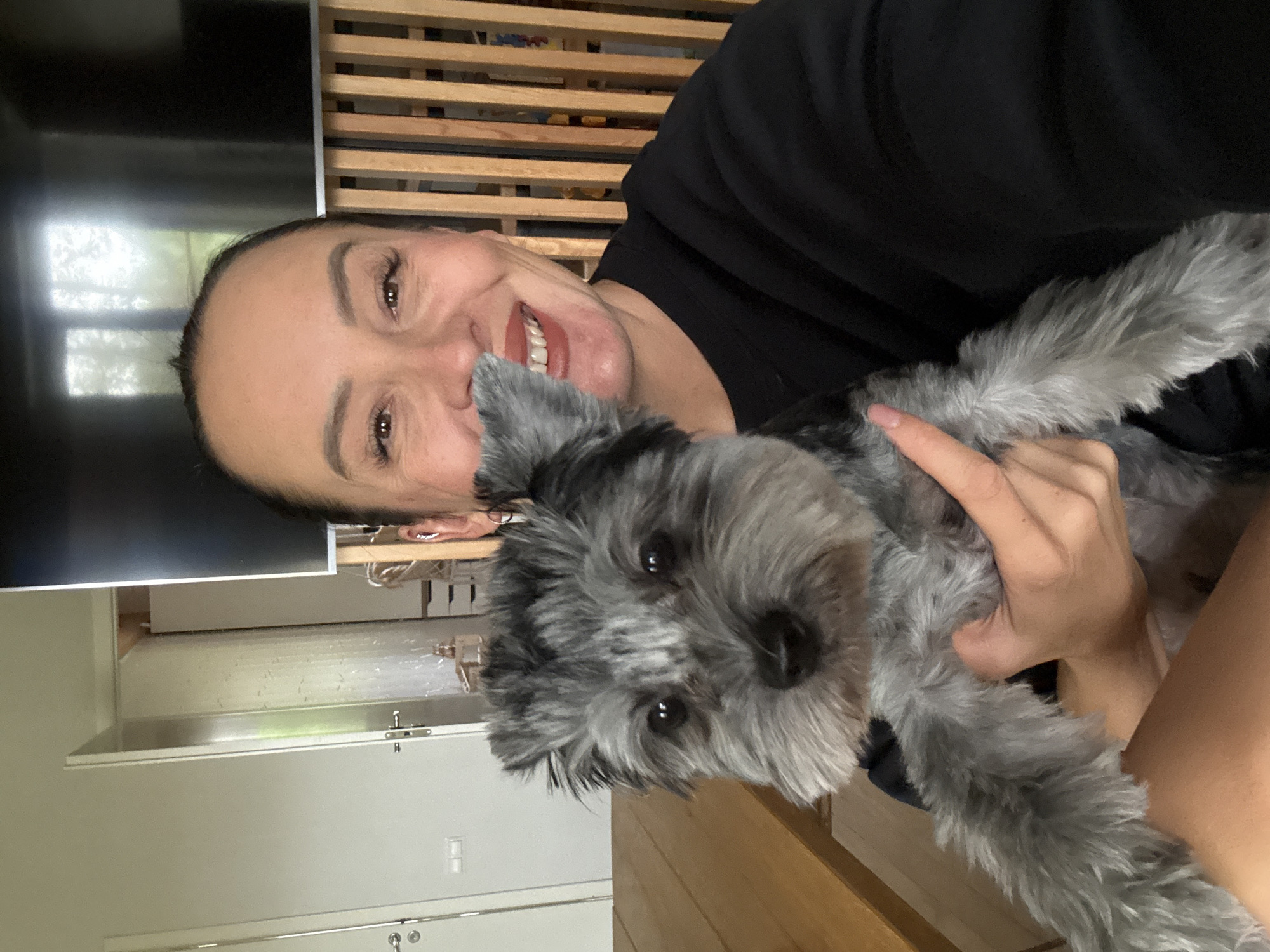 An upper body shot captures a woman with dark hair pulled back, smiling broadly at the camera while holding a small, gray-and-black dog. The woman is wearing a black sweatshirt. The dog, positioned in the foreground, has a shaggy coat and large, dark eyes. Behind them, a wooden slatted partition is visible, and to the left, a plain white door and a wooden table are present. The background suggests an indoor domestic setting, possibly an apartment or house, and the overall lighting is bright. No specific location in Chaikovsky, Russia, can be determined from the visual information.