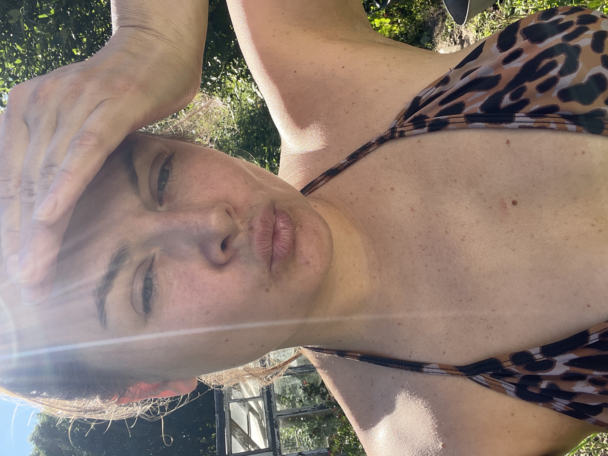 The media file is a close-up, low-angle selfie taken outdoors during the daytime. The subject is a woman wearing a leopard-print bikini top. She is holding her hand to her forehead, shielding her eyes or perhaps indicating discomfort from the sun. The lighting suggests it is a bright, sunny day, with lens flare visible in the image. In the background, there are blurred green foliage and a greenhouse structure, suggesting a garden or outdoor recreational area. The woman's expression appears somewhat pensive or perhaps a bit strained due to the bright light. There is no visible text in the image. The location context is Заречный, Russia.