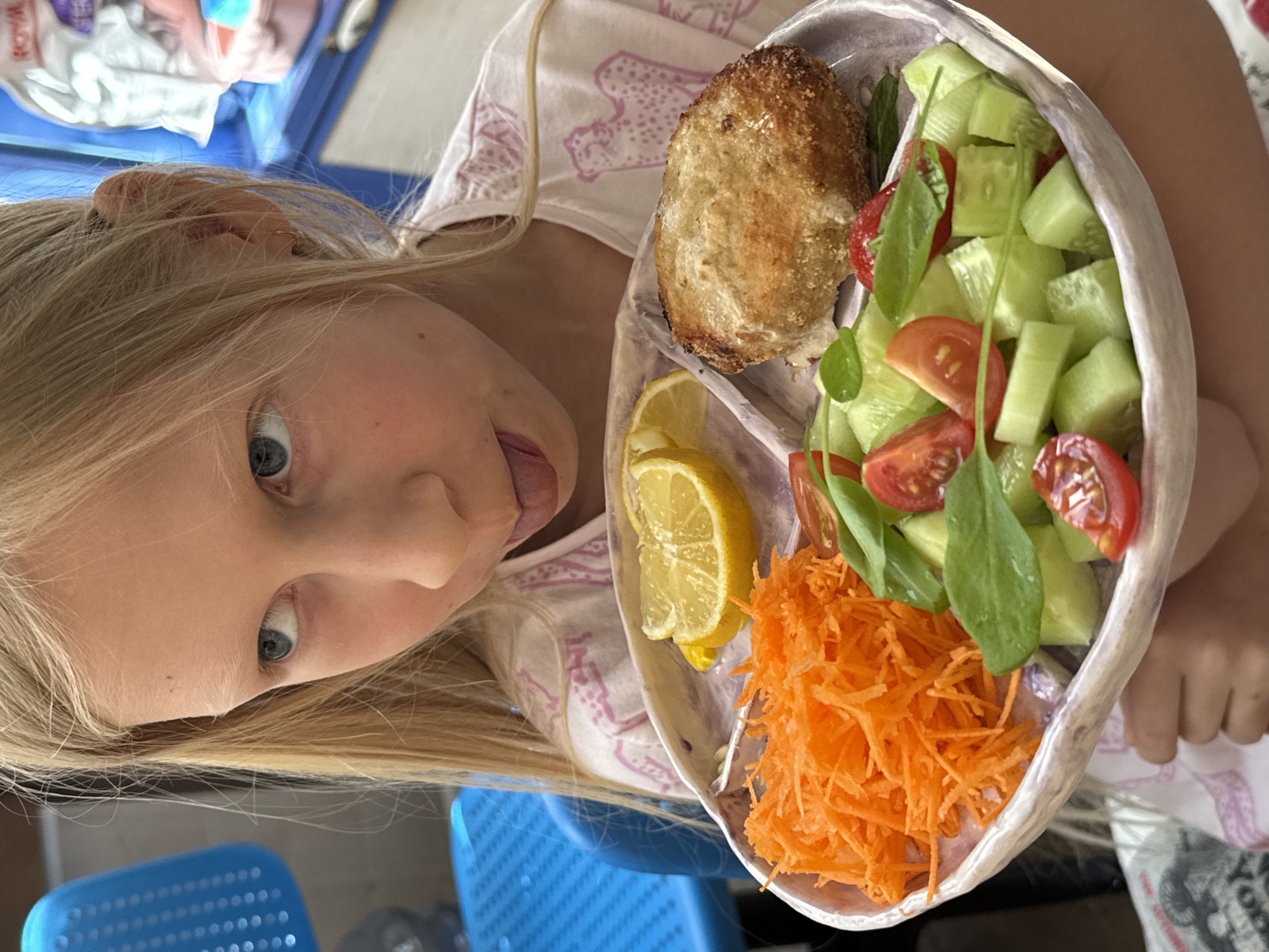 A blonde-haired child with blue eyes is holding a plate of food. The plate contains a piece of bread, two lemon slices, shredded carrots, and a cucumber tomato salad with greens. The child is sticking their tongue out, appearing playful. In the background, there is a blue plastic chair and other indiscernible objects. There is no information to indicate that the photo was taken in Chaikovsky, Russia.