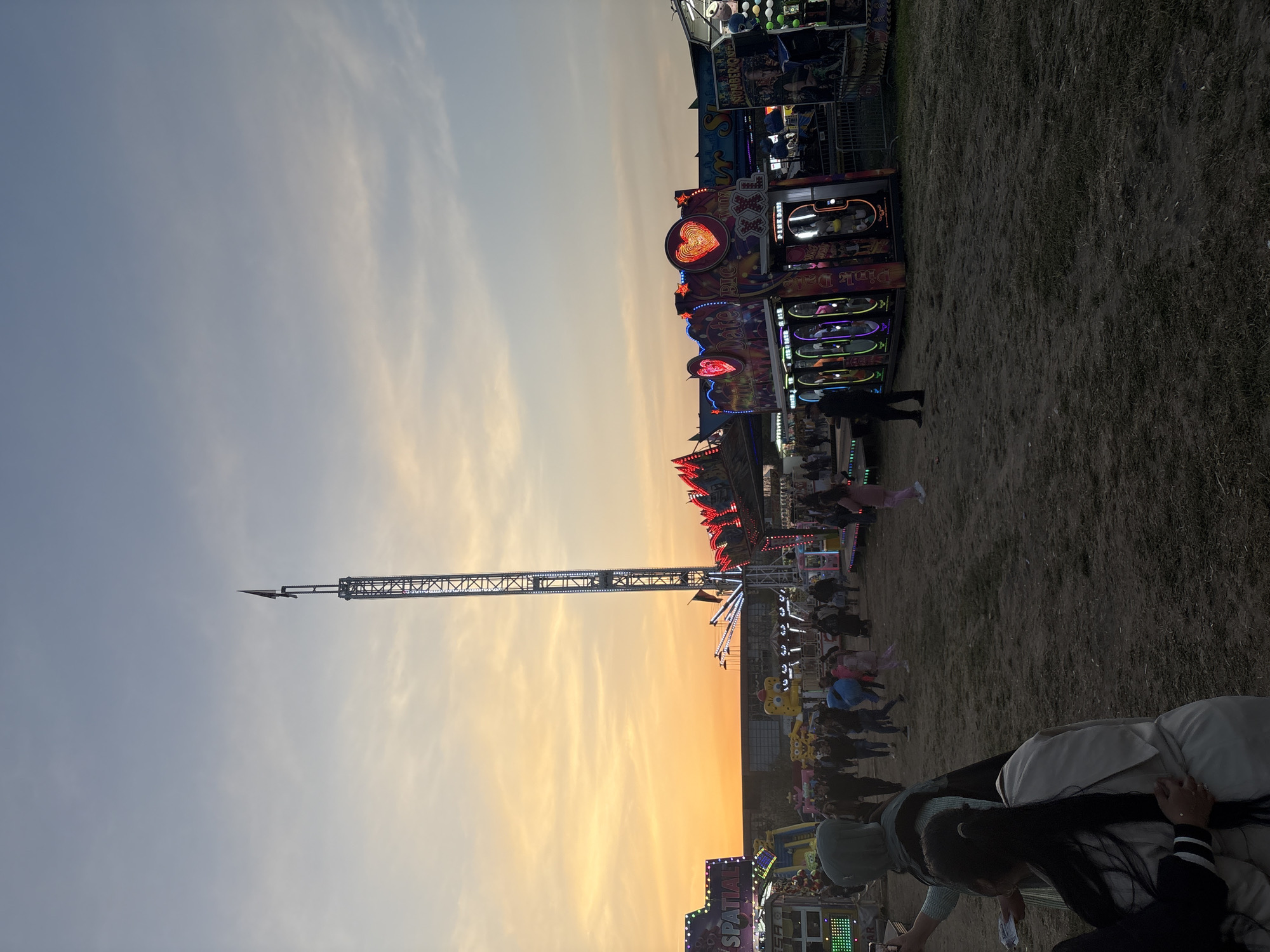 The image shows a fairground at dusk. A tall tower ride dominates the skyline, reaching into the orange-tinted sky. Carnival games and attractions line the ground, illuminated with colorful lights. People are milling about, though they are not clearly visible due to the dim lighting. The ground appears to be covered in grass or dirt. The location is Unknown, Unknown.
